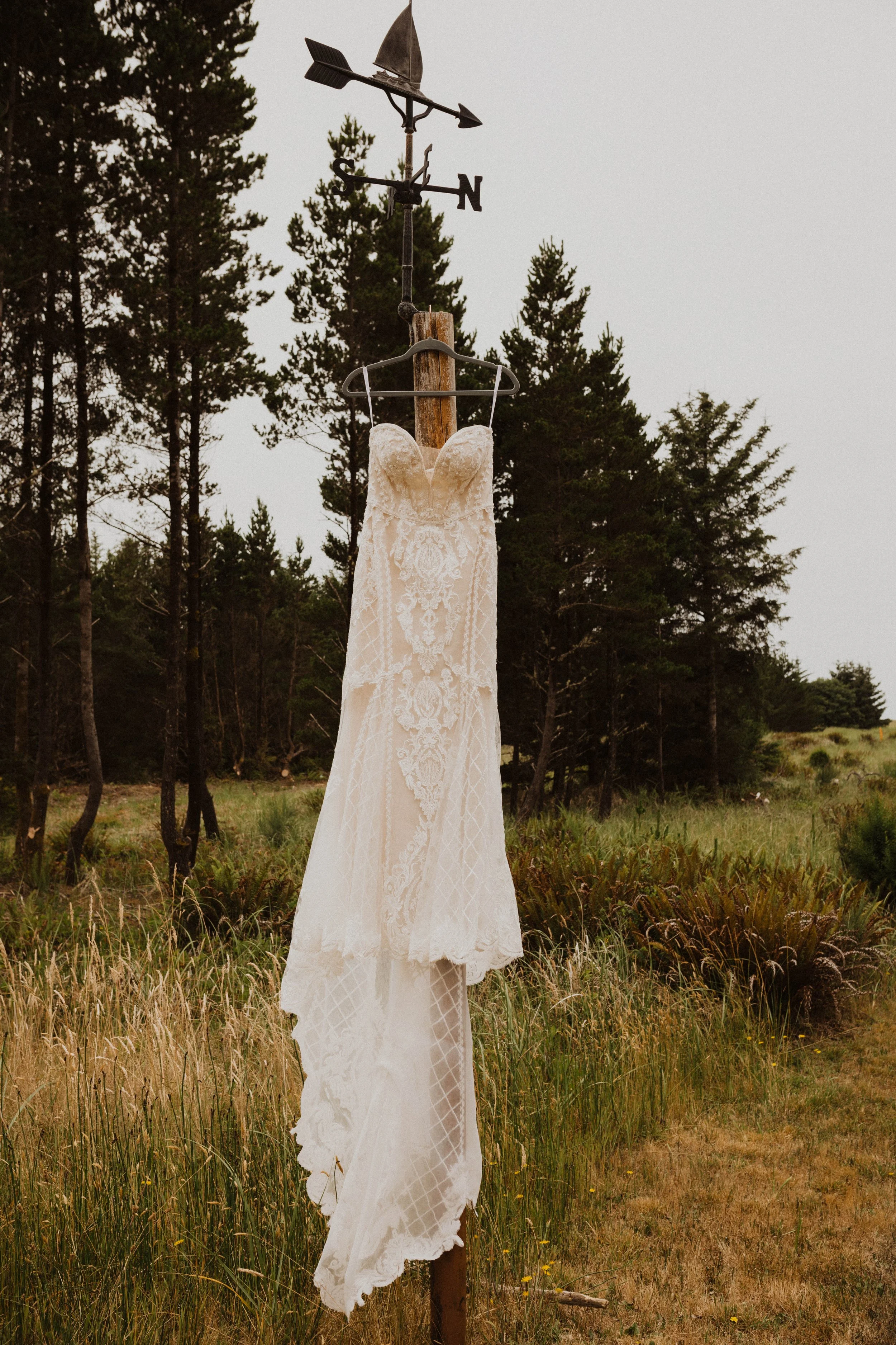 A wedding dress hanging from a weather vane on a wooden pole in an outdoor natural setting with trees and grass. Long Beach, WA wedding photography.