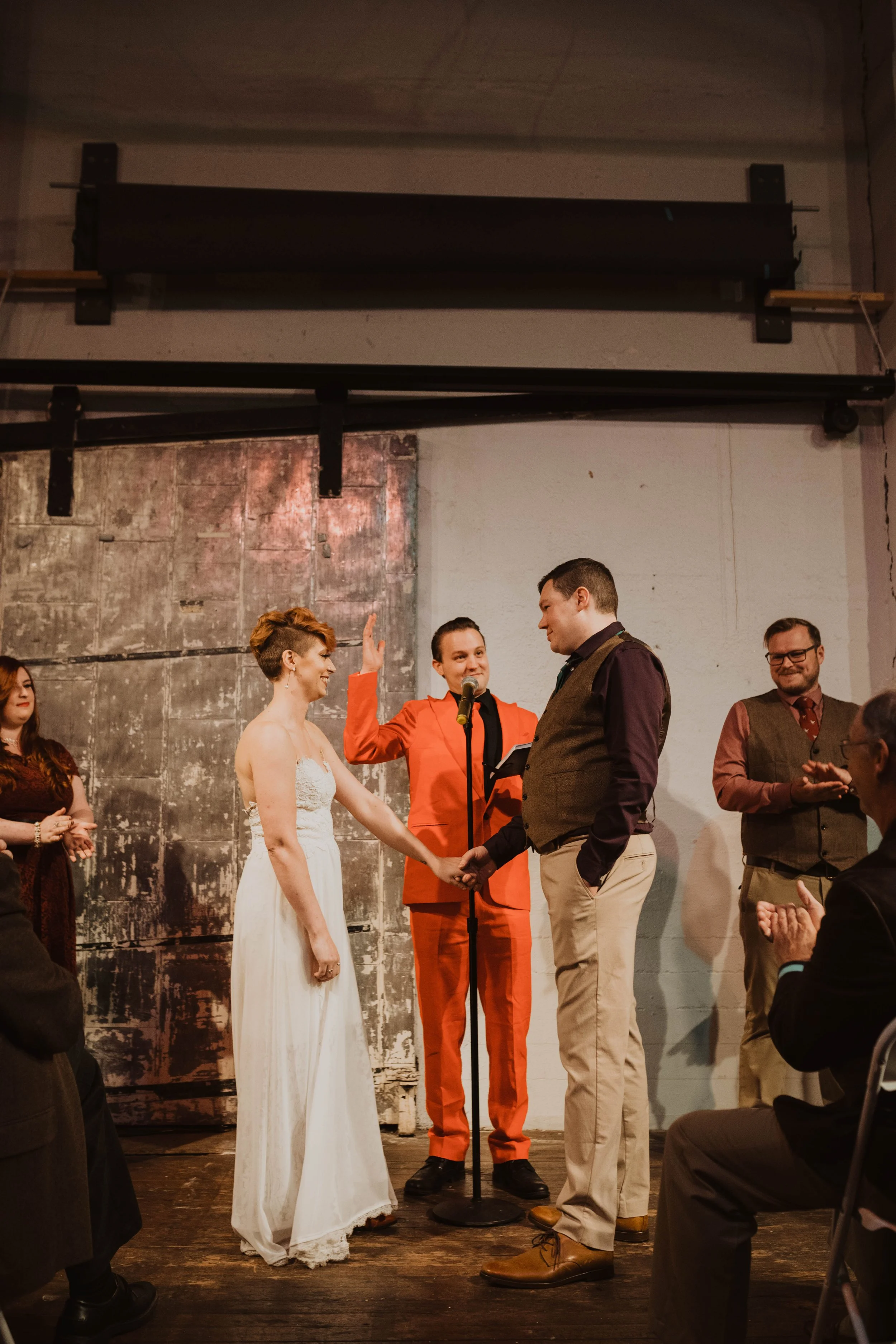 A wedding ceremony with a bride and groom holding hands, an officiant raising his hand, and guests clapping in a rustic industrial venue. Pioneer Square, Seattle, WA wedding photography.