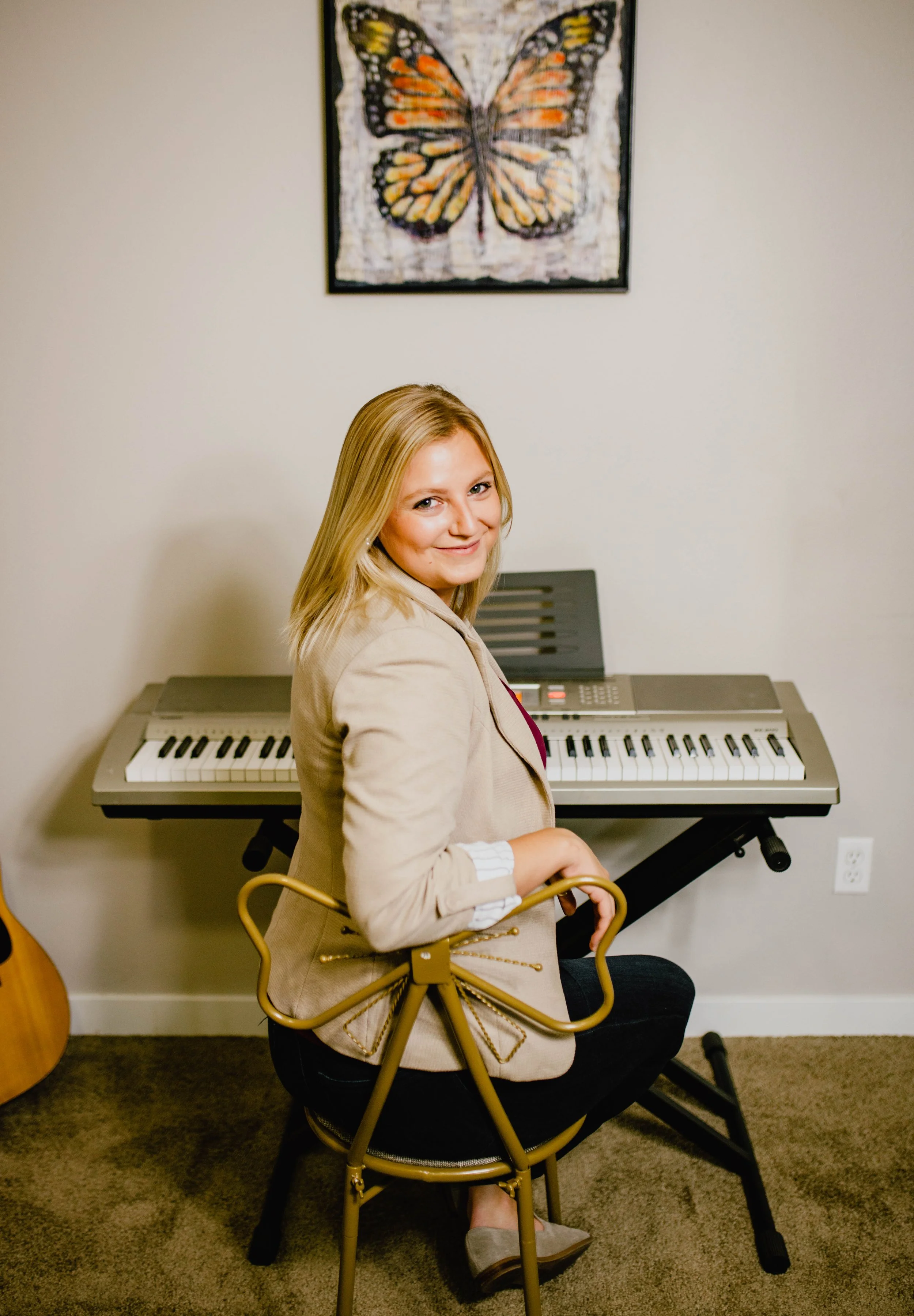 A woman with blonde hair sitting on a black chair in front of a digital keyboard, smiling at the camera, in a room with beige walls and a butterfly artwork hanging above the keyboard. Seattle professional head shot photography