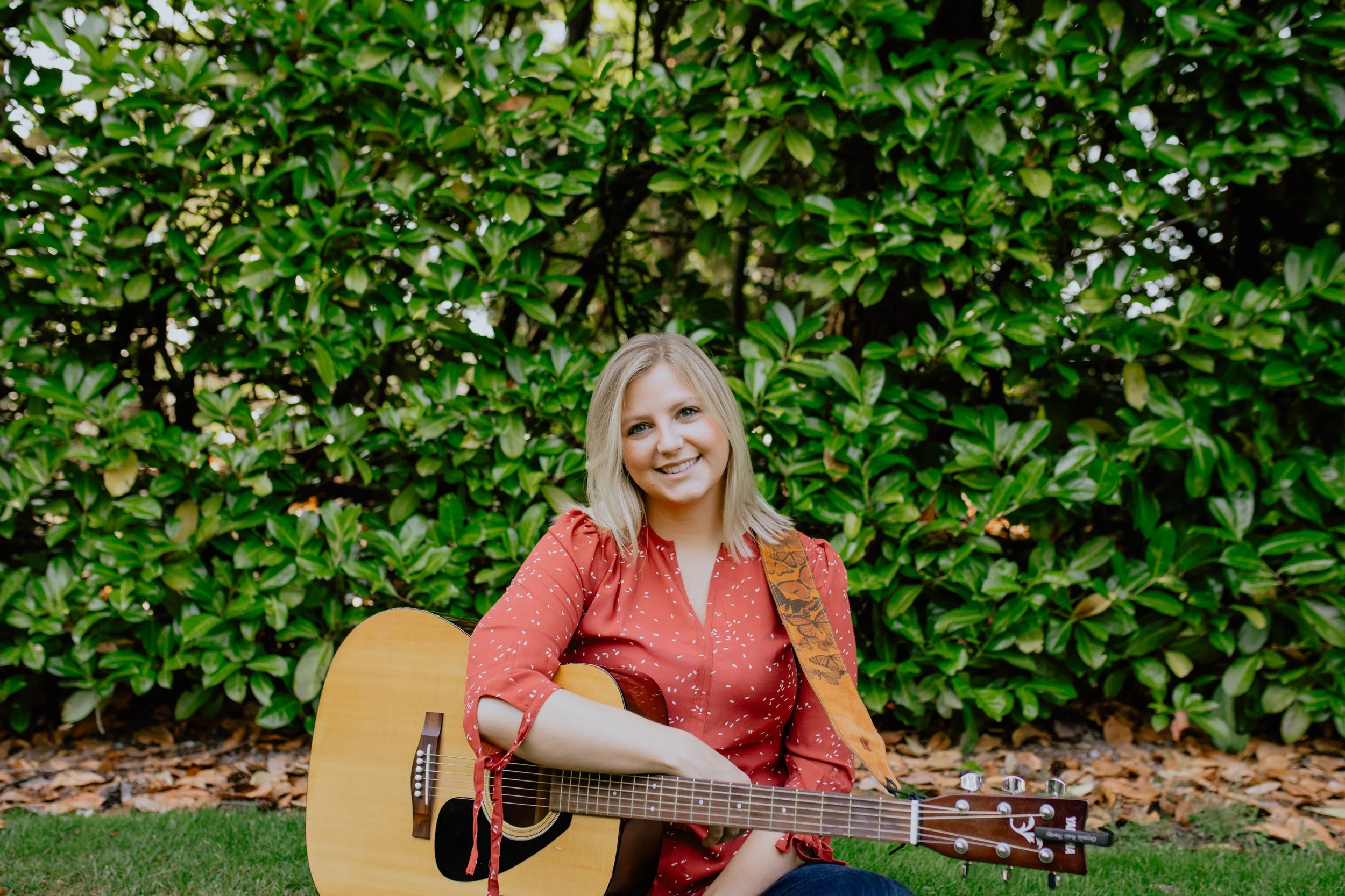 A woman with blonde hair sitting on grass in front of green bushes, holding an acoustic guitar and smiling. Seattle professional head shot photography