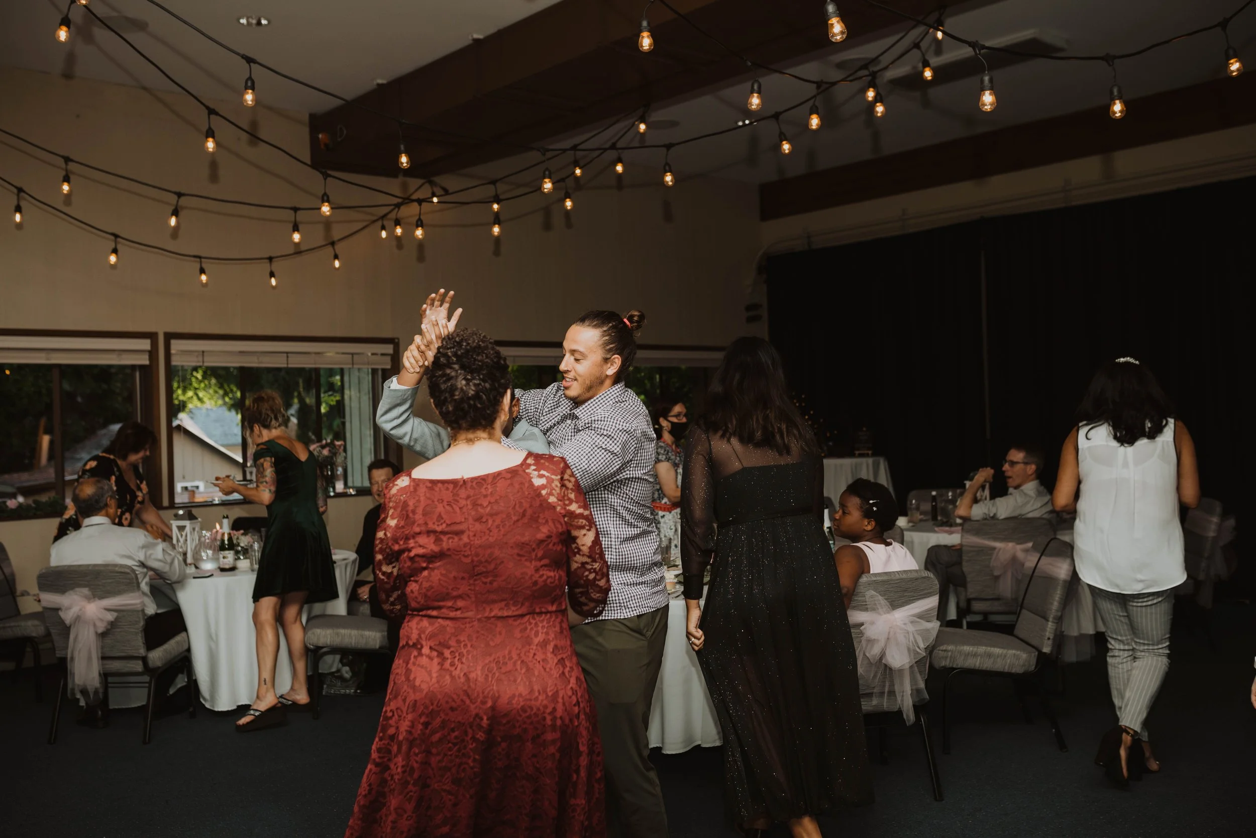 People dancing and socializing at an indoor event with string lights hanging from the ceiling, decorated tables with chairs, and windows showing greenery outside. Seattle, WA wedding photography.