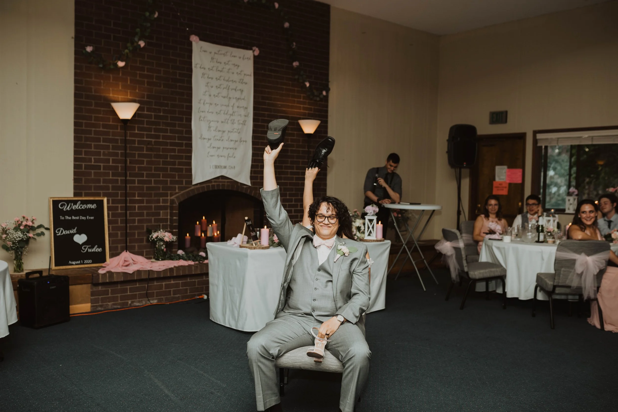 A groom in gray suit with a pink bow tie, glasses, and a boutonniere, sitting on a chair and holding black shoes in the air at a wedding reception. The reception is decorated with flowers, candles, and a sign with the couple's names, David and Tundee