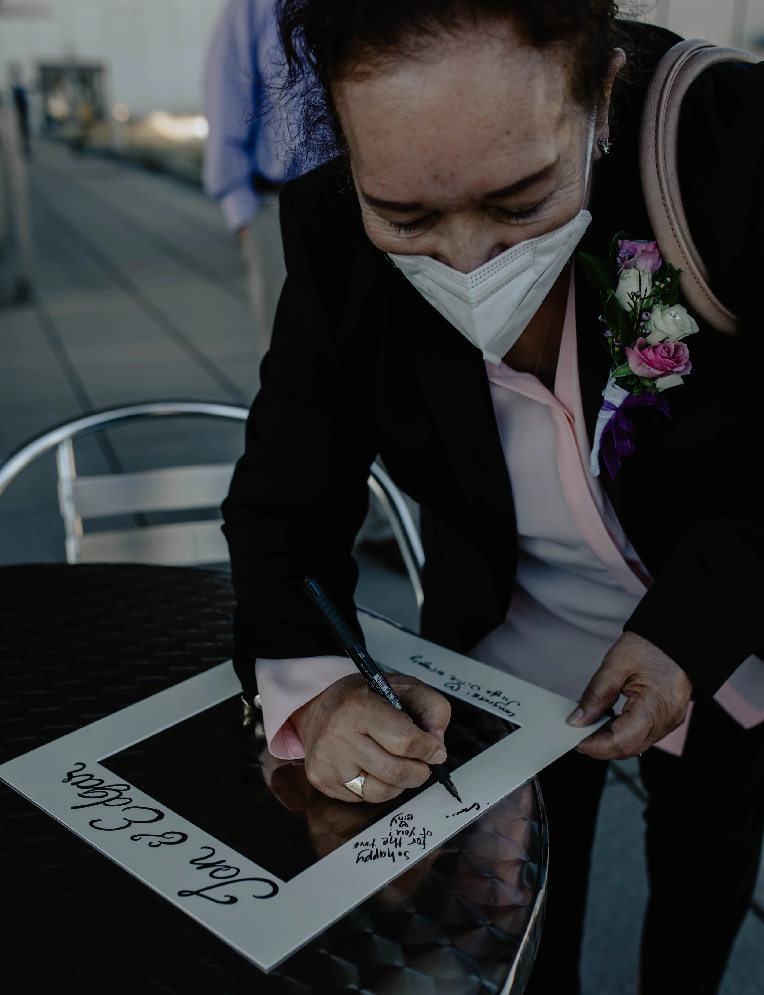 Woman in a black jacket and pink shirt signing a framed photograph or poster with a black marker, wearing a white face mask and a flower corsage, outdoors on a patio with a blurred background. Seattle Municipal Courthouse wedding photography.