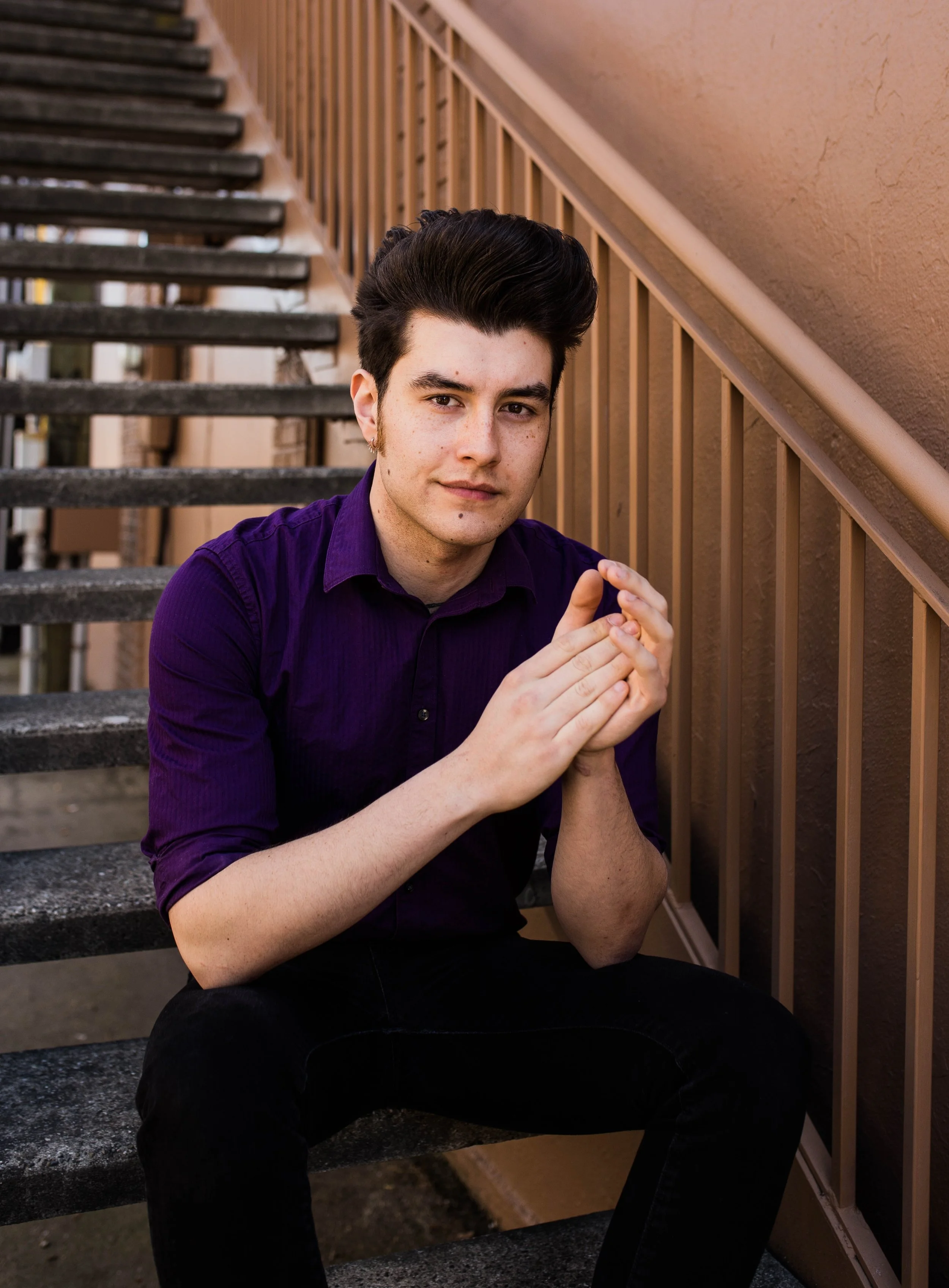 A young man with dark, styled hair and light skin, sitting on outdoor stairs with a tan railing, wearing a purple button-up shirt and black pants, looking at the camera with a slight smile. Seattle professional head shot photography