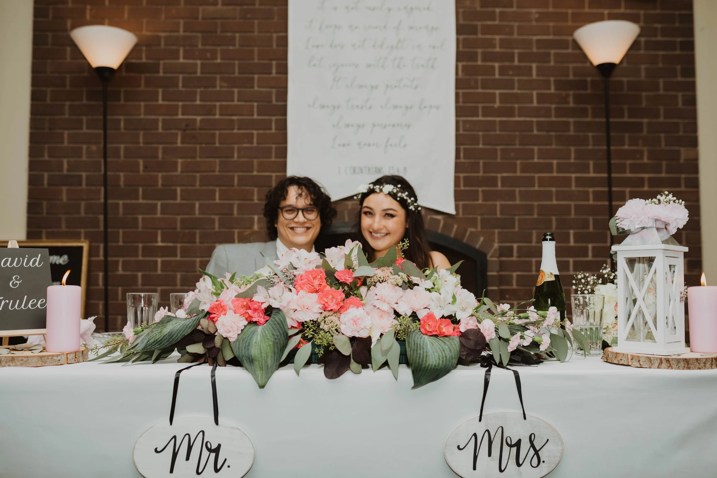 Happy bride and groom sitting at a decorated wedding table with pink and white flowers, candles, and a white lantern, against a brick wall background with wall sconces and a hanging banner. Seattle, WA wedding photography.