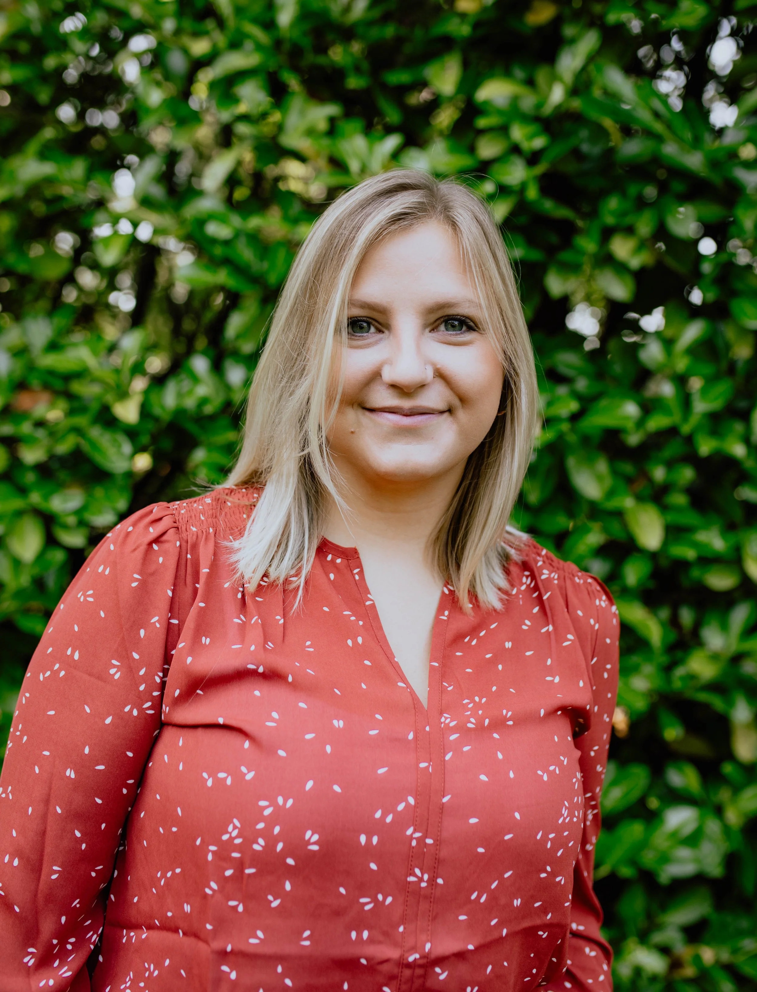 A woman with blonde hair and blue eyes, smiling, wearing a red blouse with white spots, standing outdoors in front of green foliage. Seattle professional head shot photography