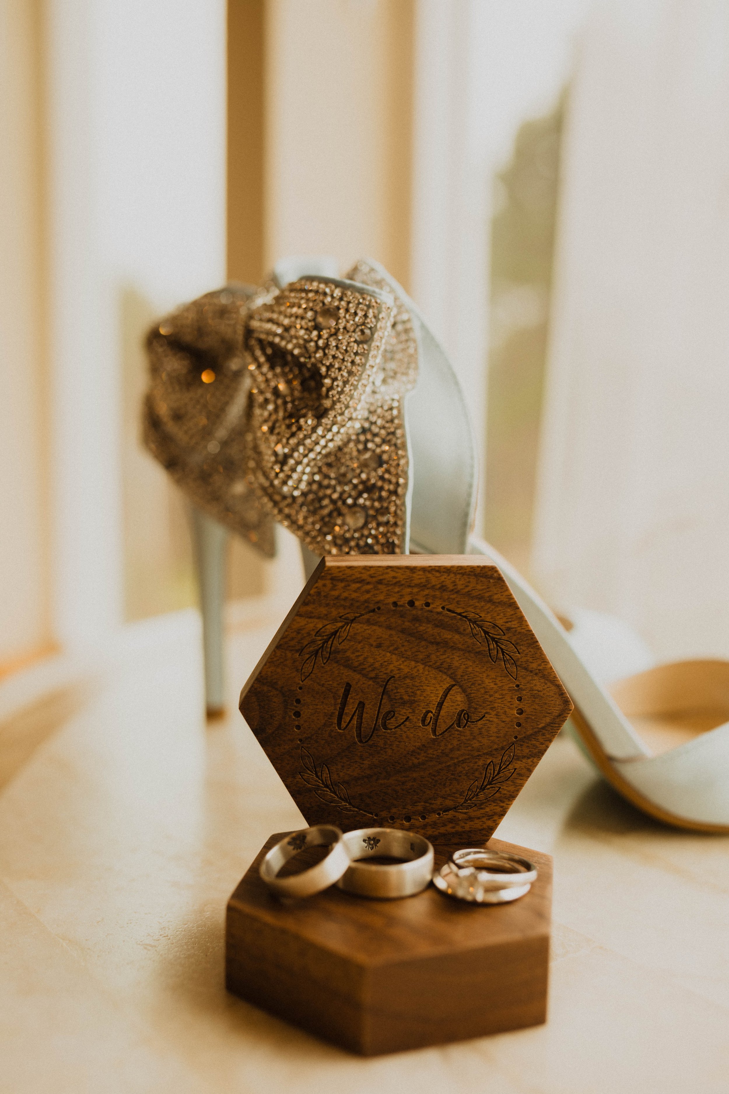 Wedding rings on a wooden display next to a hexagonal wooden sign with 'We do' engraved on it, with a high-heeled shoe with rhinestones and a silver brooch behind. Long Beach, WA wedding photography.