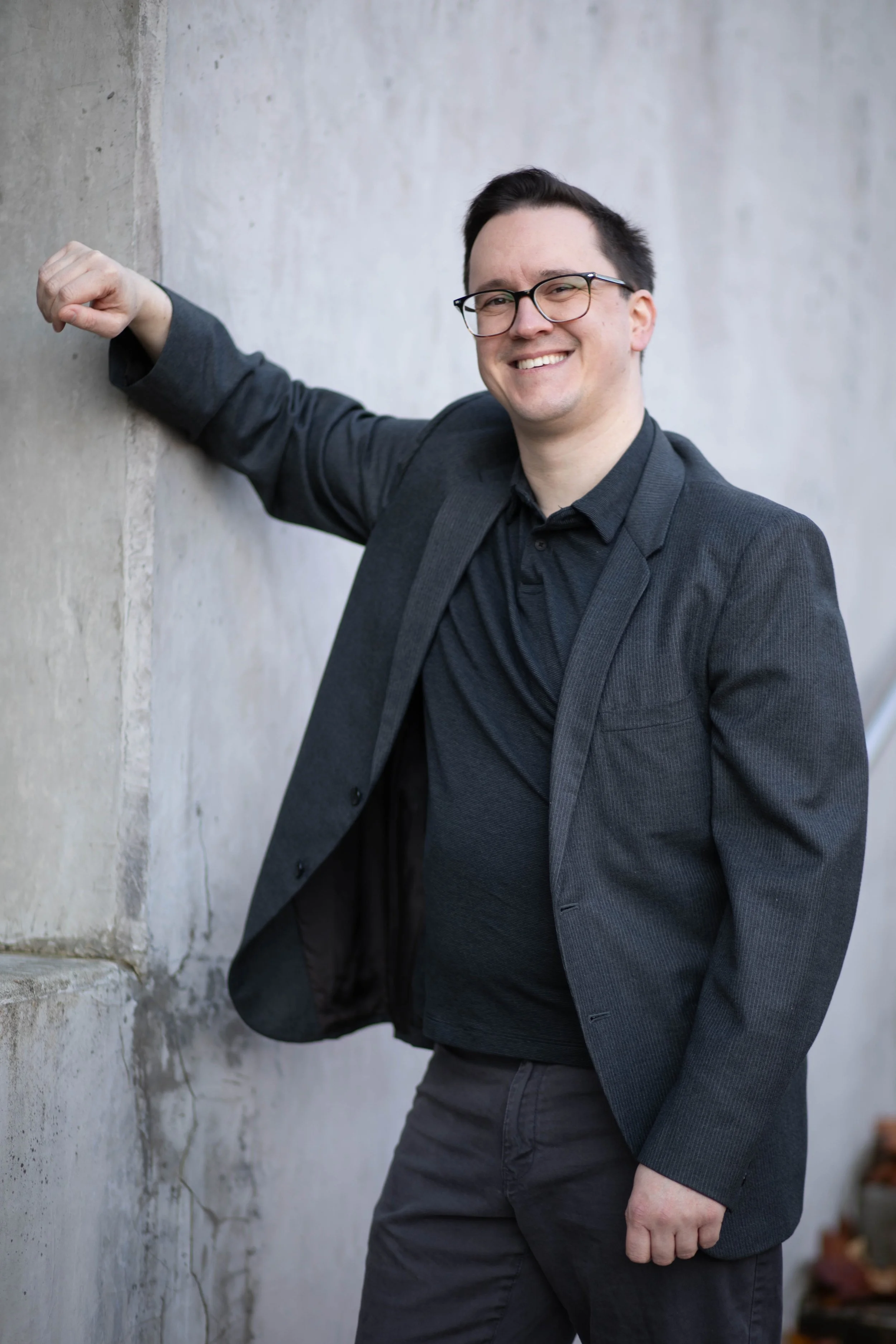 A man in a dark blazer and glasses smiling, leaning against a concrete wall with one arm resting on it. Seattle professional head shot photography