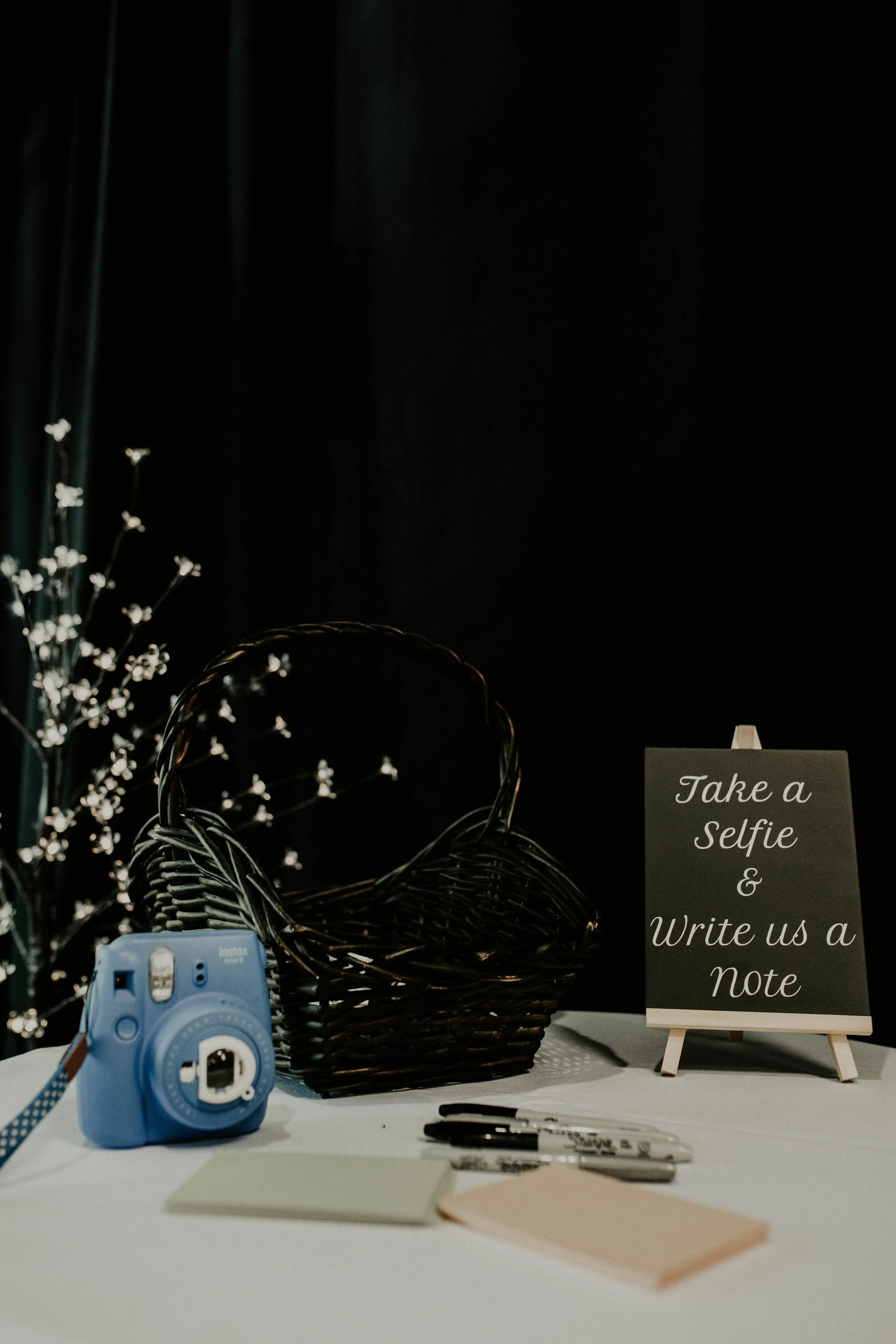 A table with a sign that says 'Take a Selfie & Write us a Note,' a blue instant camera, black and white markers, sticky notes, and a black wicker basket on it. In the background, there is a dark curtain and a decorative branch with lights. Seattle, W