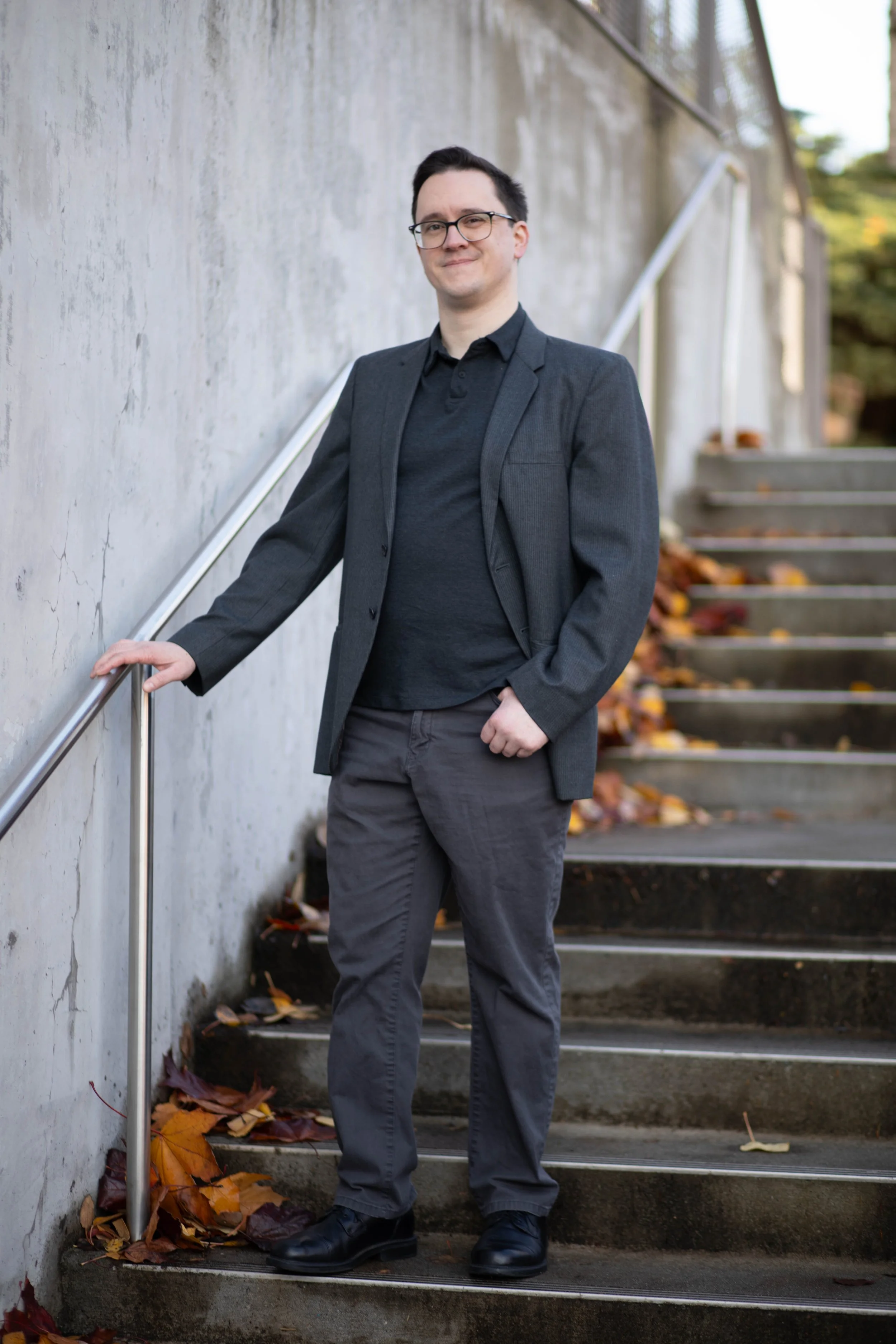 A man wearing glasses, a dark blazer, and dark pants standing on outdoor concrete stairs with autumn leaves, leaning on a metal handrail near a textured concrete wall. Seattle professional head shot photography