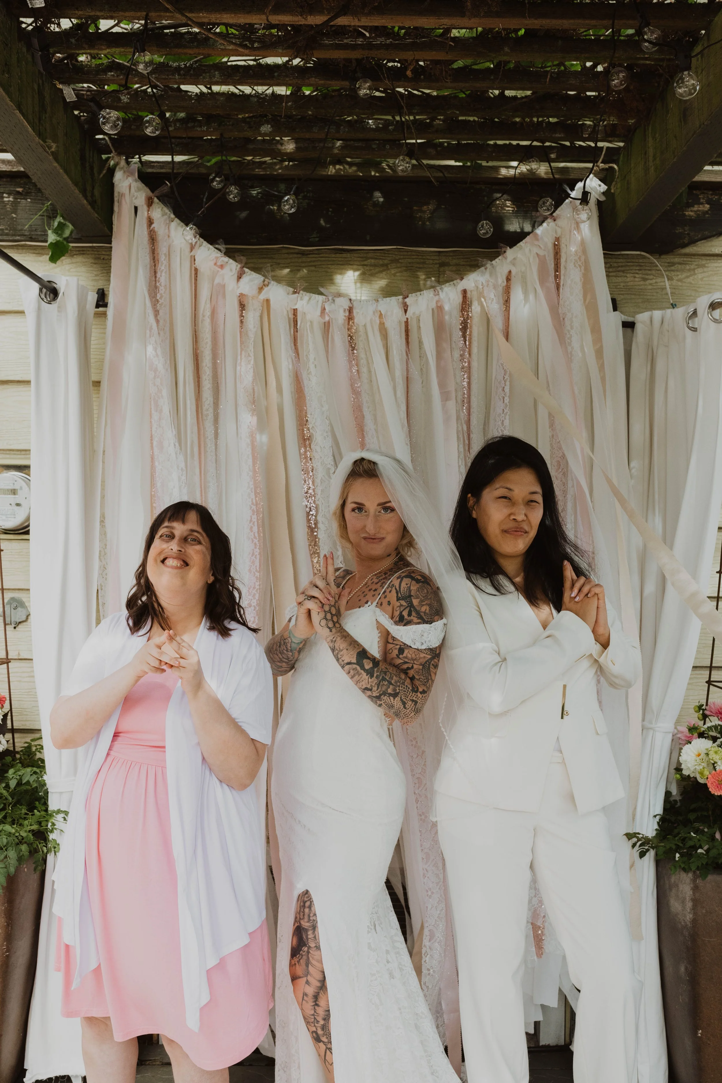 Three women posing together, one in a wedding dress with tattoos, standing in front of a pastel-colored curtain backdrop, outdoors. Seattle, WA wedding photography.