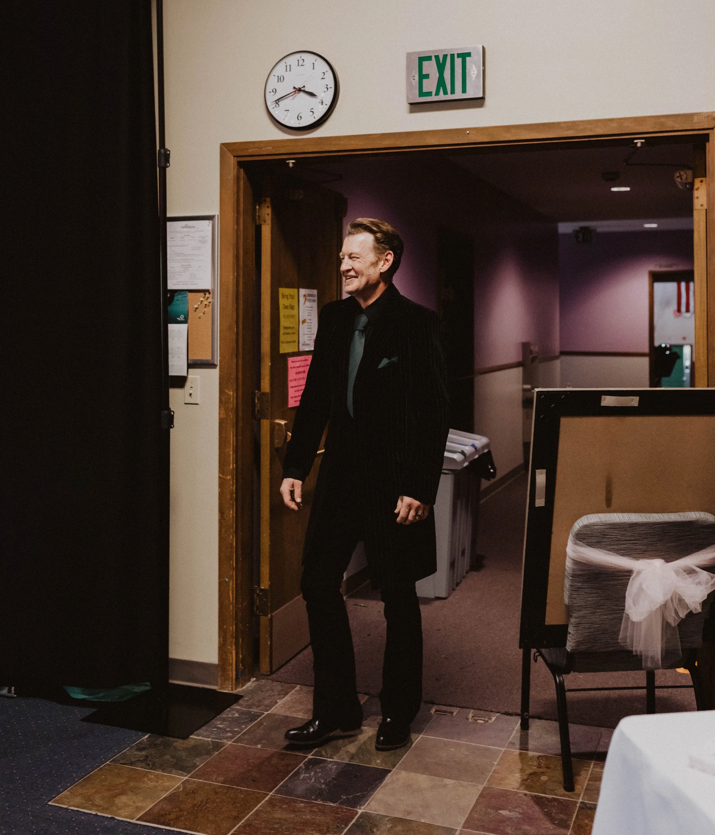 A man in a black suit with a teal tie smiling as he stands in a doorway inside a building, with an 'EXIT' sign and a clock showing 10:43 behind him. Seattle, WA wedding photography.