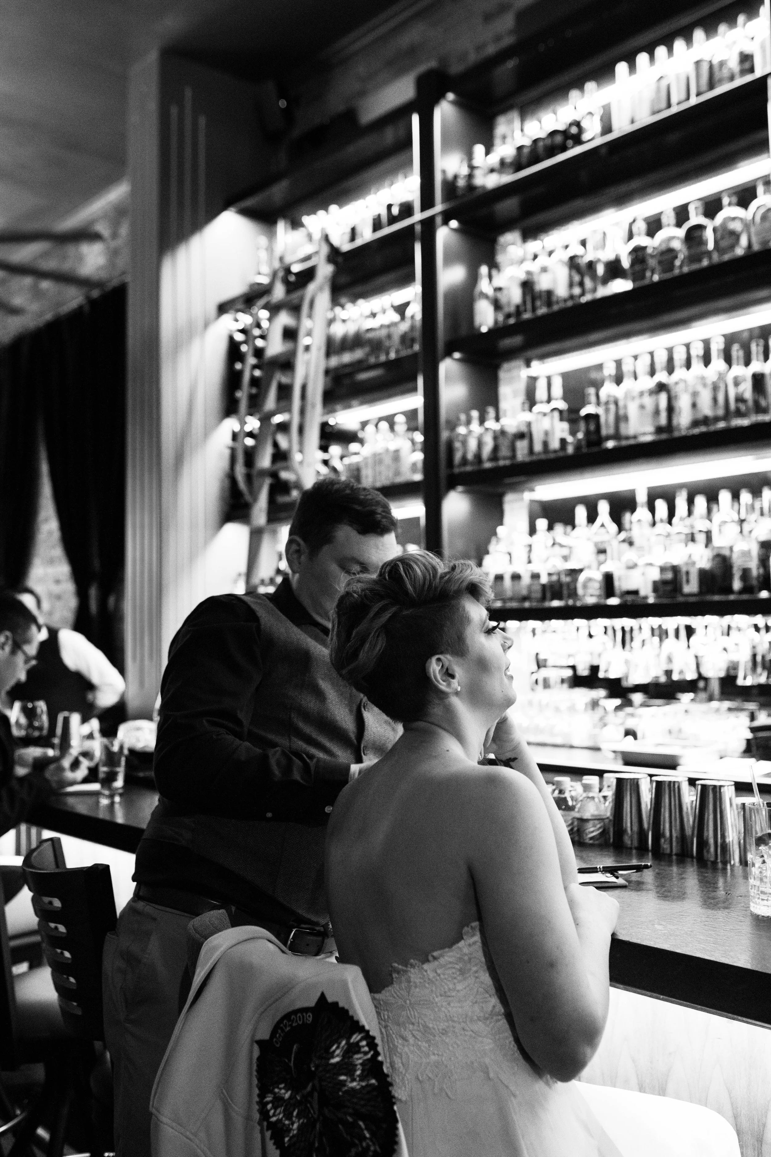 A woman in a strapless dress sitting at a bar with a man standing behind her, in a dimly lit bar or restaurant, with shelves of bottles behind them. Pioneer Square, Seattle, WA wedding photography.