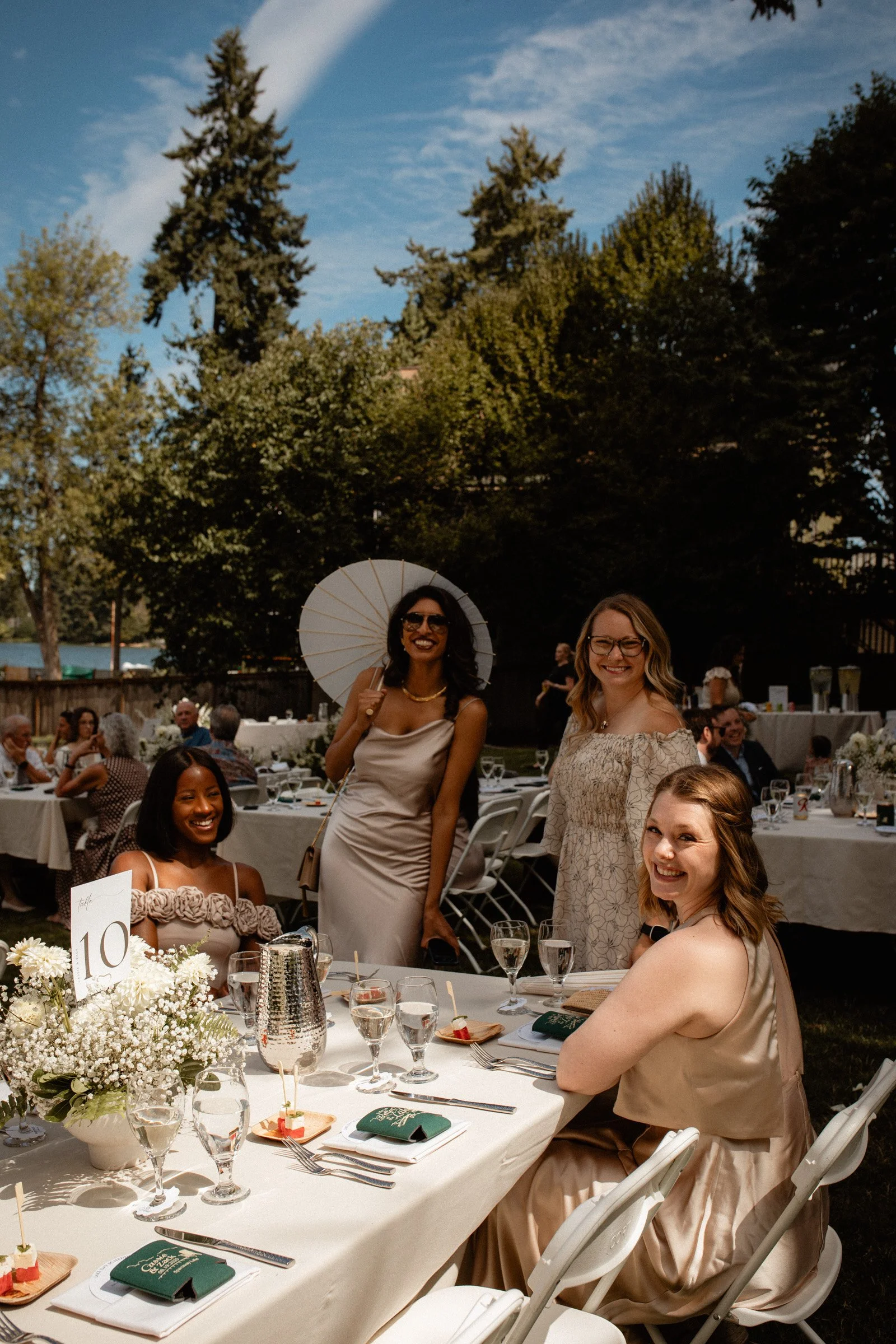 Wedding guests mingle at a table together holding umbrellas for shade at this lakeside wedding in Seattle, WA