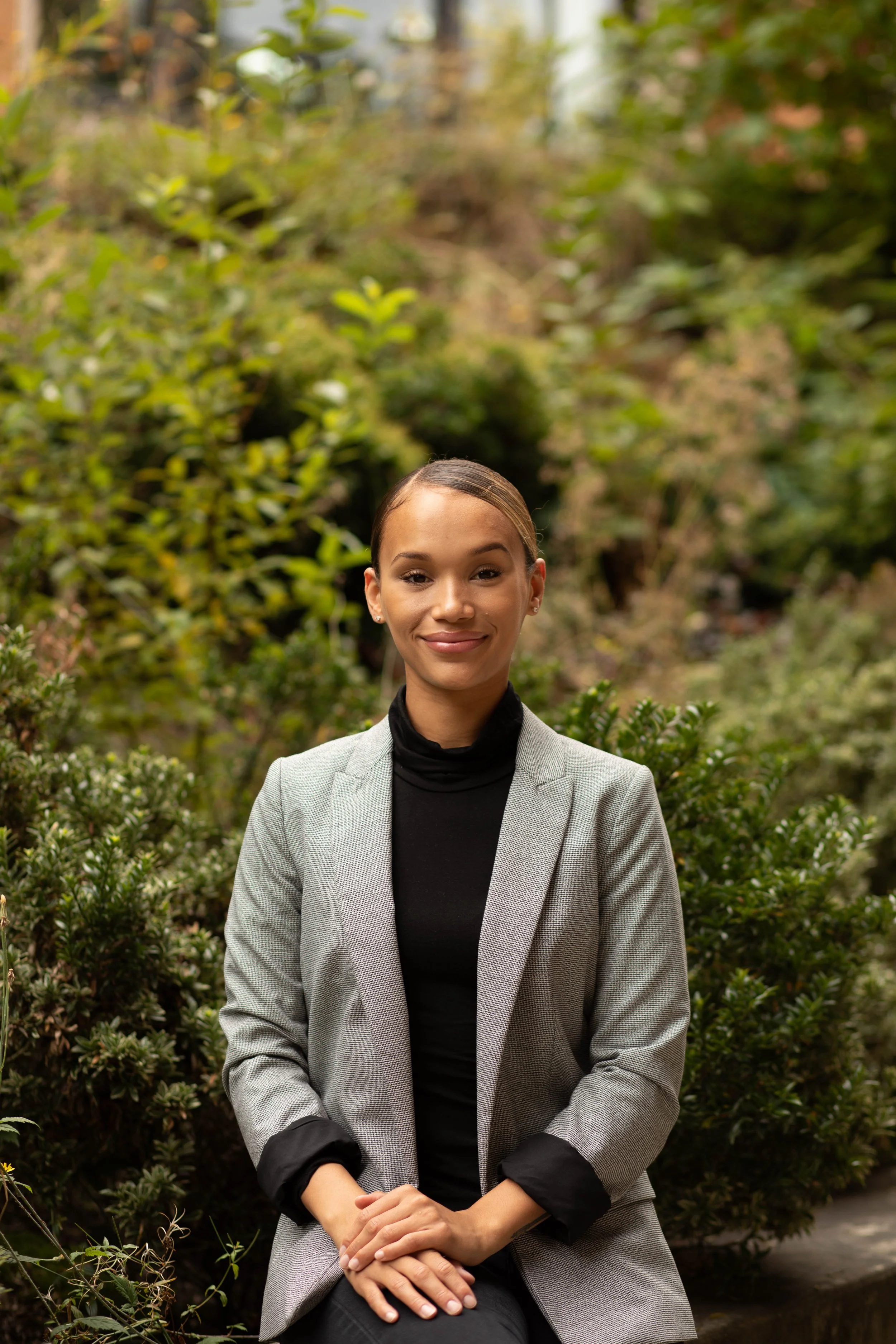 A young woman with a warm smile, sitting outdoors in a lush garden, wearing a gray blazer over a black shirt. Seattle professional head shot photography