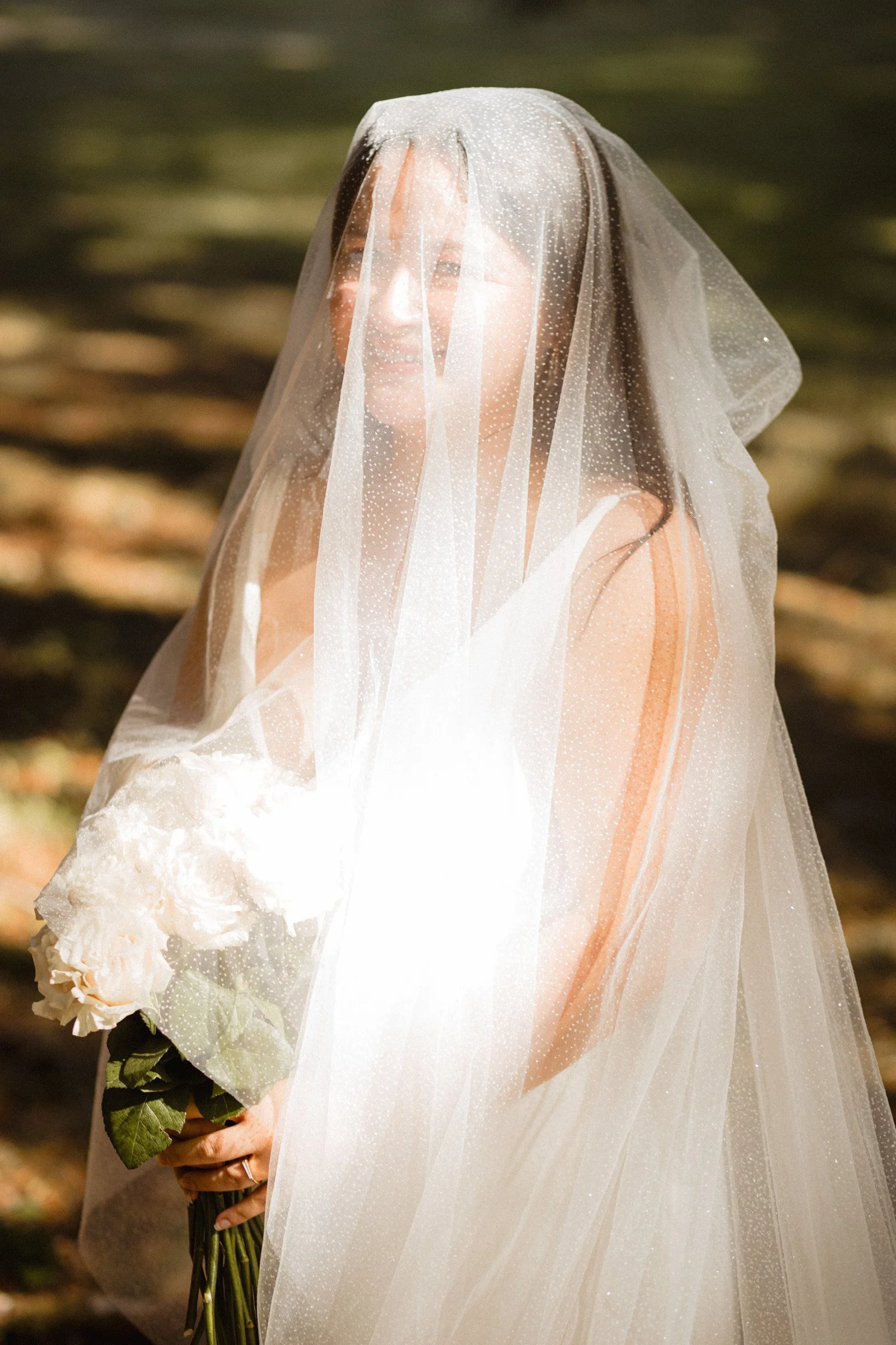 A bride stands smiling at the camera with her veil drapped over her and her simple white bouquet. She stands in the forest moments before she walks down the aisle in Lake Crescent, Port Angeles, WA.