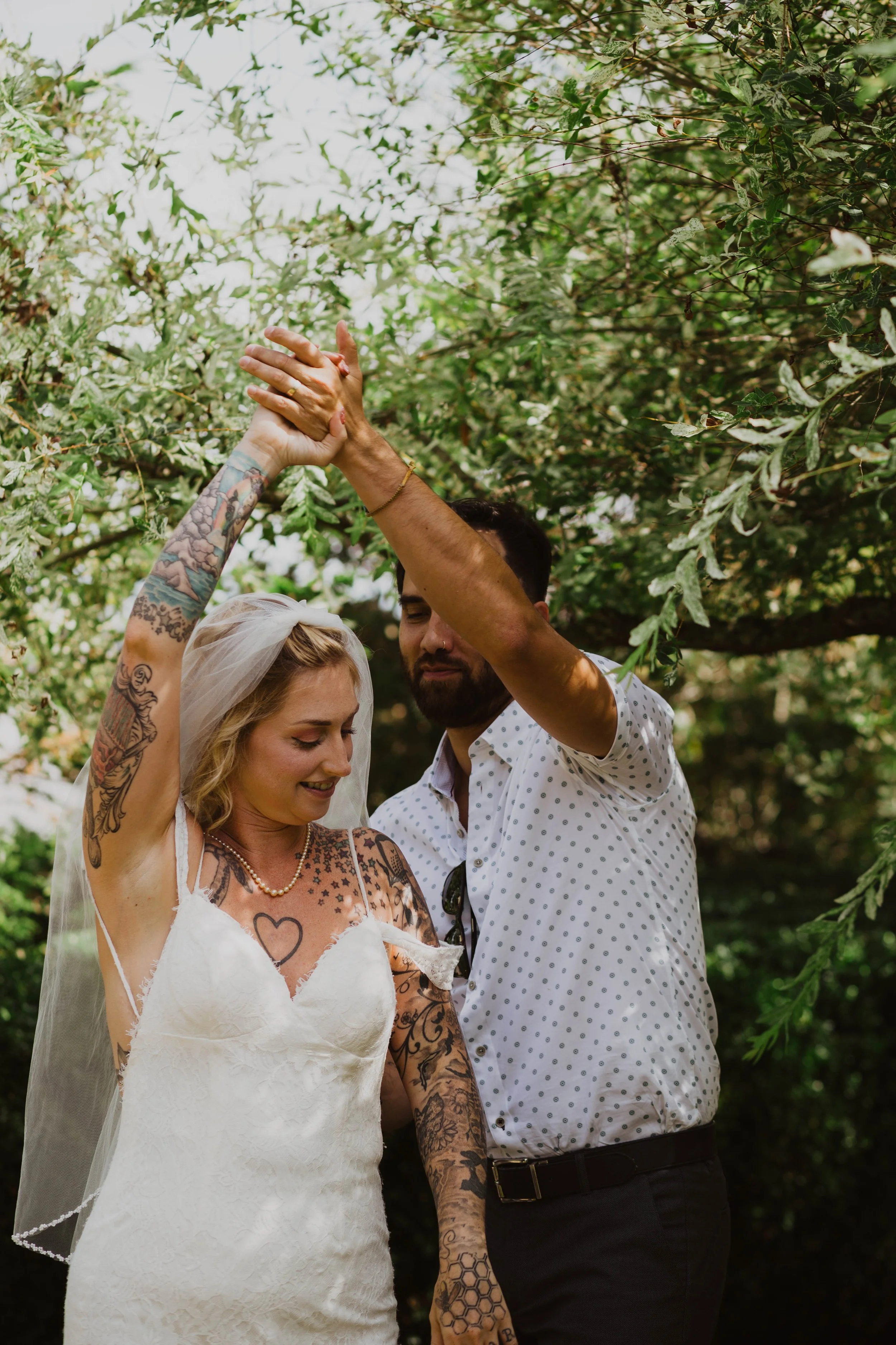 A woman and a man are dancing outdoors under a canopy of green leaves, with the woman wearing a wedding dress and veil, and the man wearing a white shirt with small polka dots. They appear to be celebrating a special occasion, possibly a wedding or a