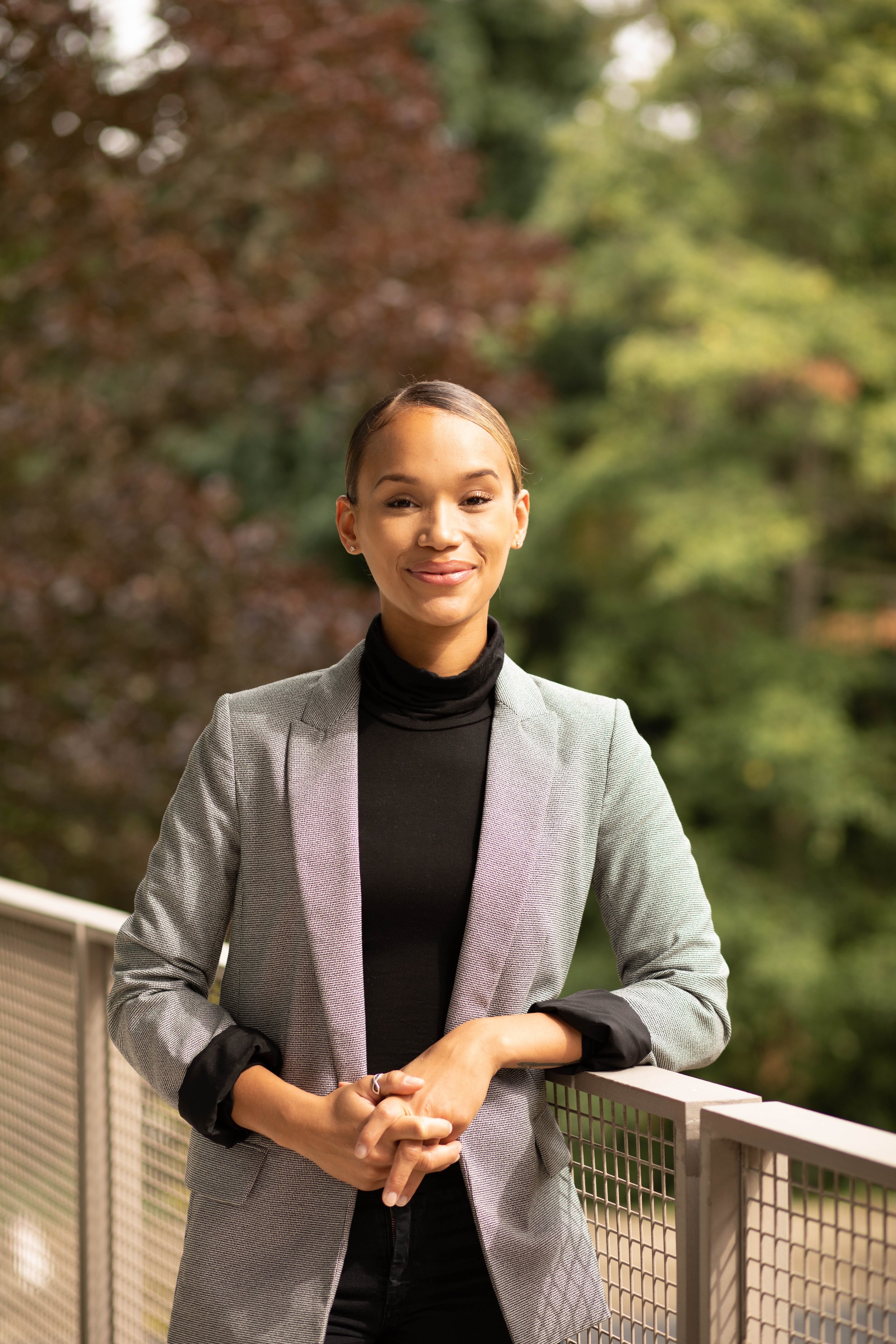 A young woman standing outdoors on a bridge, smiling with her arms crossed, wearing a gray blazer and black turtleneck, with trees and greenery in the background. Seattle professional head shot photography