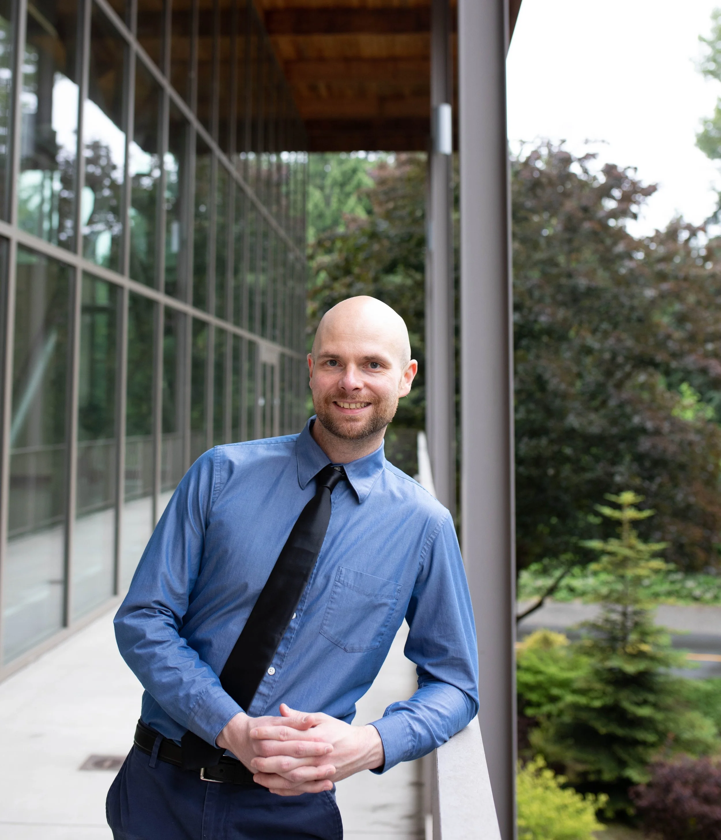 Young man with a shaved head wearing a blue dress shirt and black tie, standing outdoors on a balcony, smiling with hands clasped in front, with glass building and trees in the background. Seattle professional head shot photography