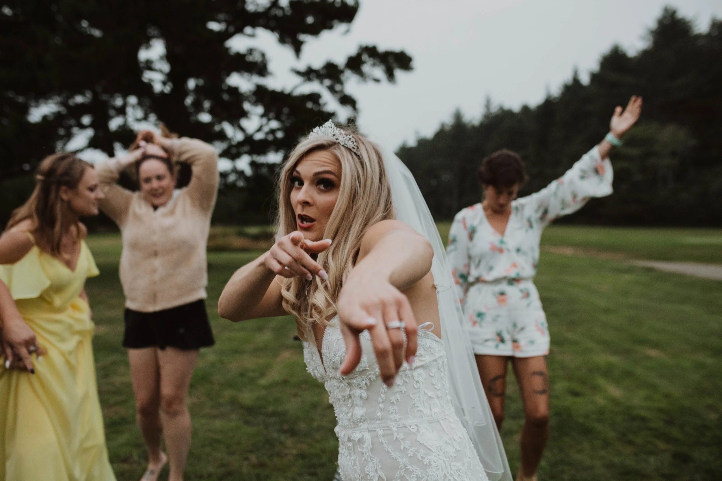 Bride in wedding dress pointing at camera, four women in casual attire dancing or posing outdoors on a grassy area with trees in the background. Long Beach, WA wedding photography.