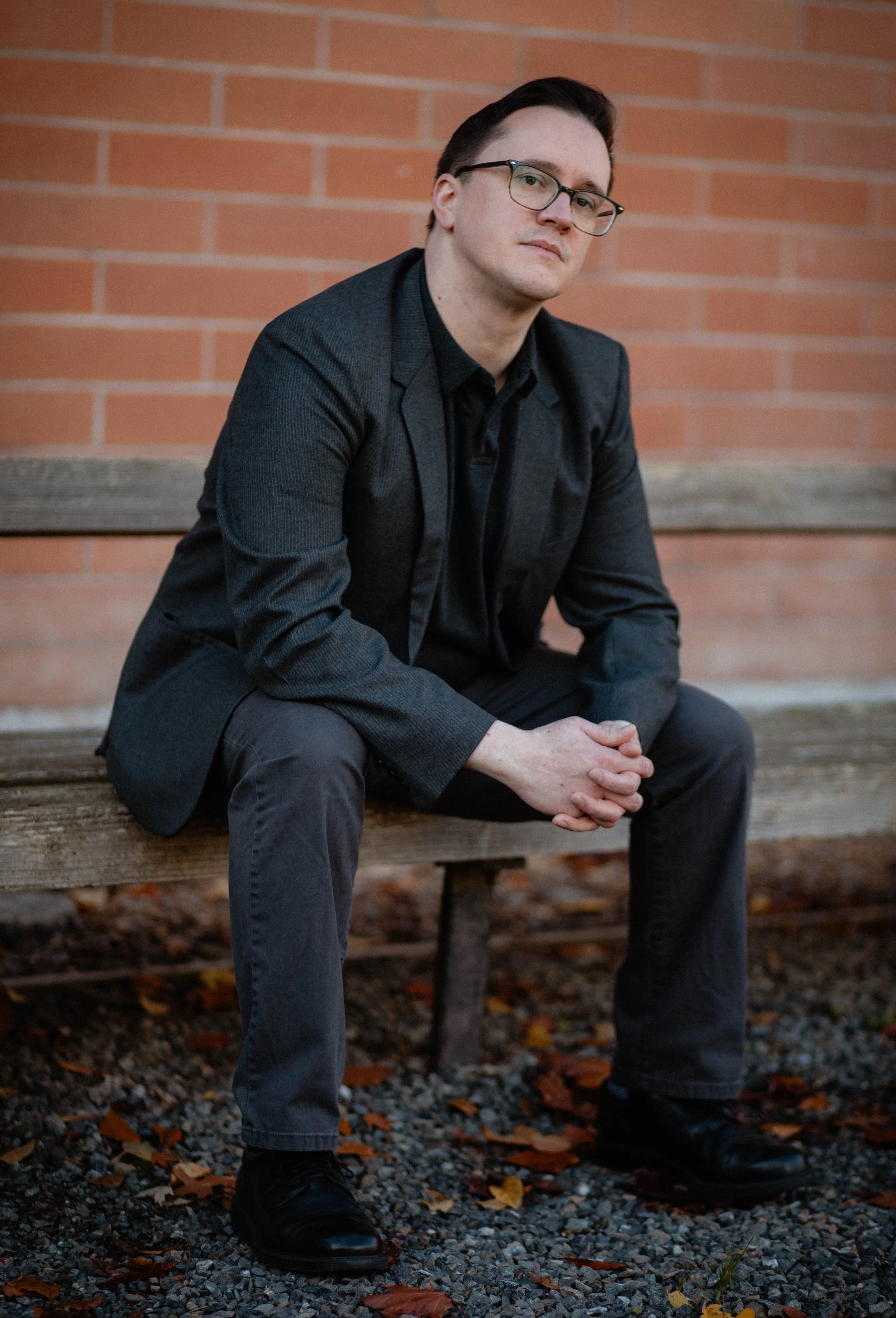 A man wearing glasses, a dark blazer, and dark pants sitting on a wooden bench with a brick wall in the background. Seattle professional head shot photography