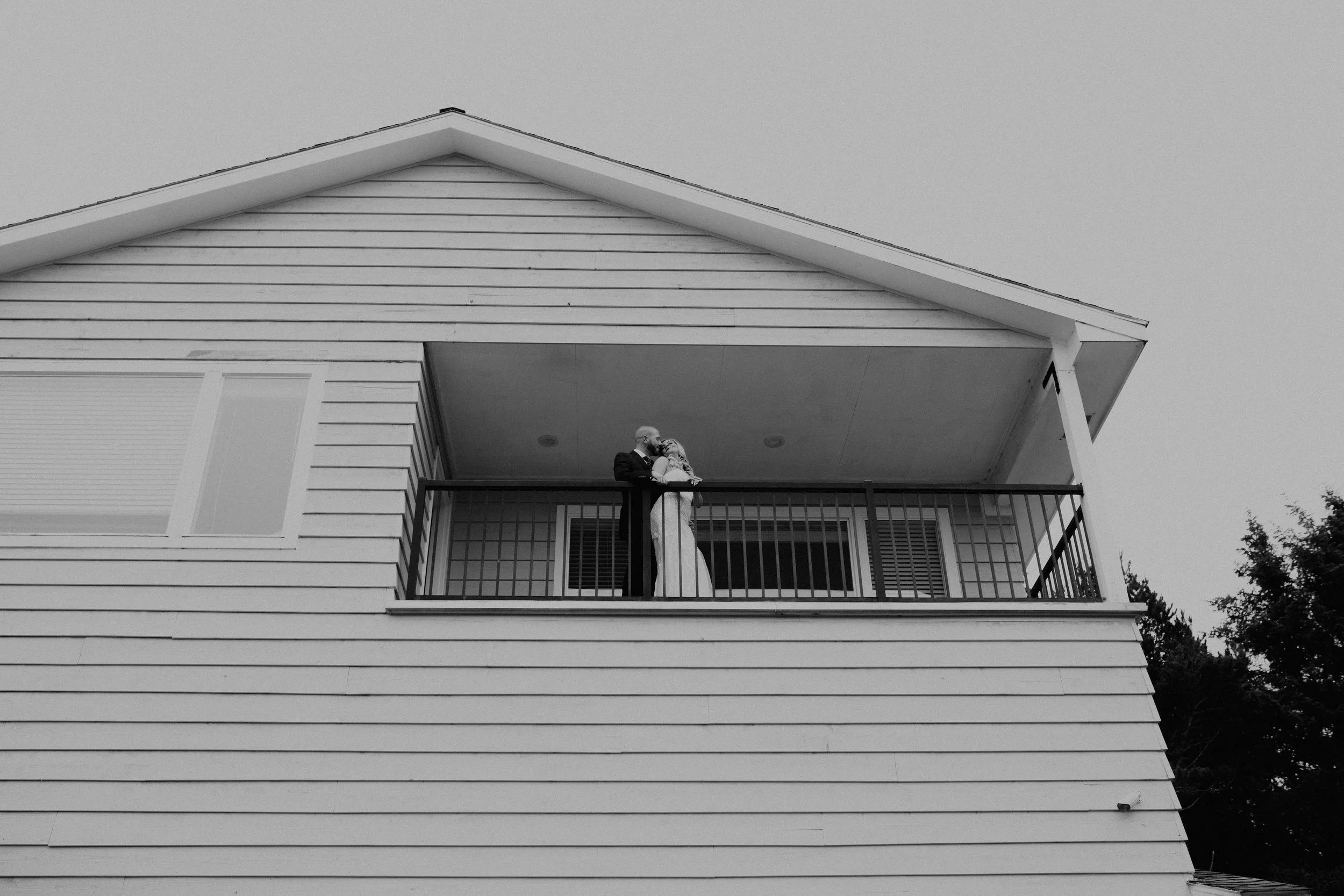 A couple in wedding attire sharing a kiss on a balcony of a two-story house with horizontal siding, seen from below. Long Beach, WA wedding photography.