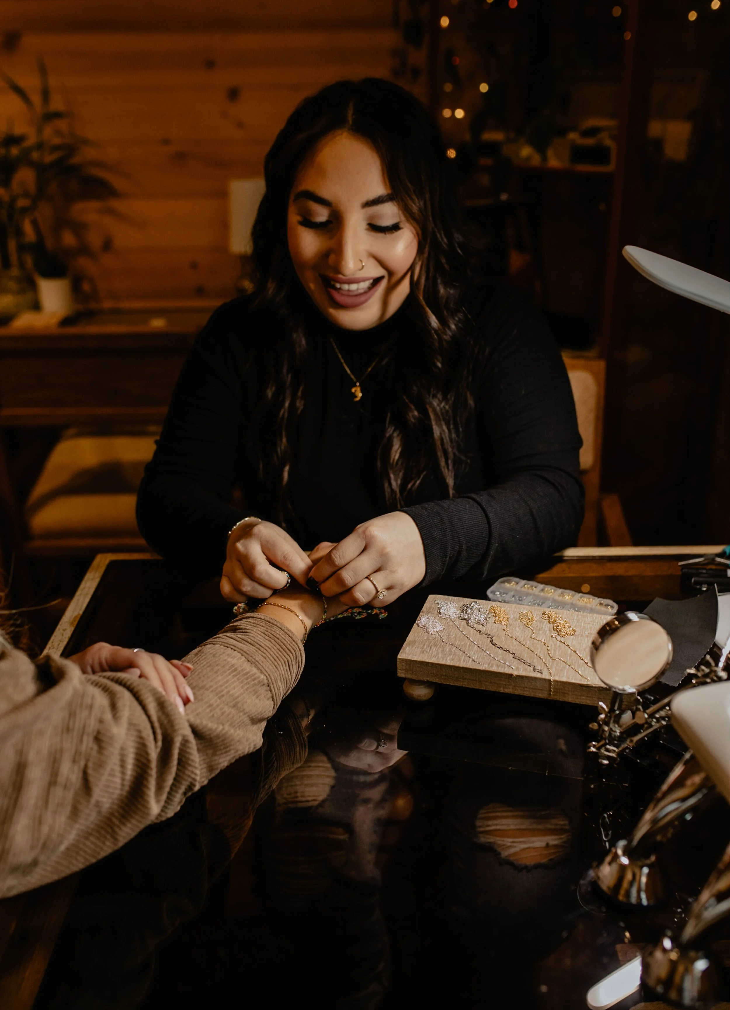 A woman attending to another person at a jewelry making or beading station, with beads and tools on the table, in a warmly lit, cozy wooden interior. Seattle professional head shot photography