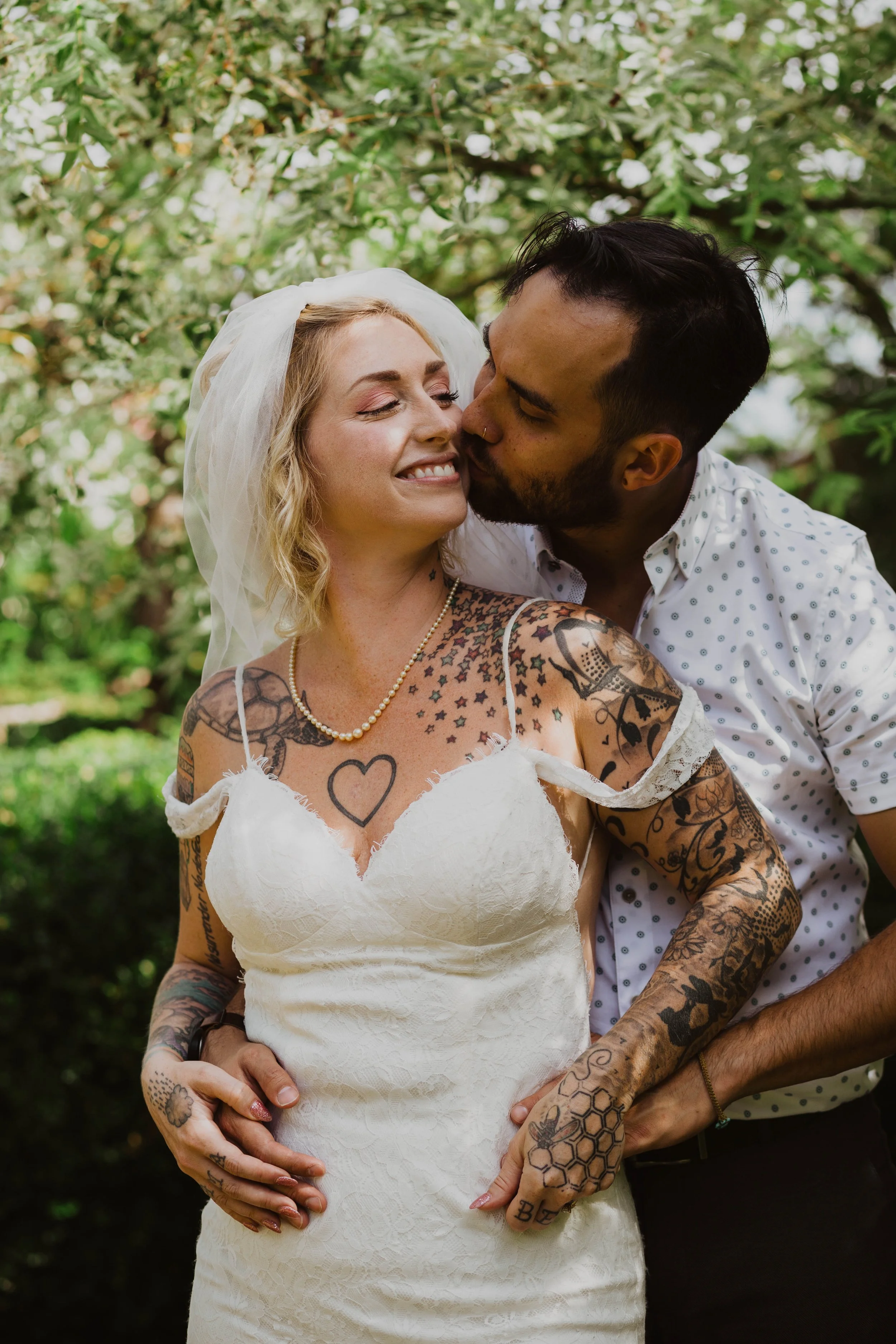 A newlywed couple sharing a kiss outdoors. The bride has blonde hair, tattoos, and is wearing a white dress and pearl necklace. The groom is kissing her cheek. Seattle, WA wedding photography.