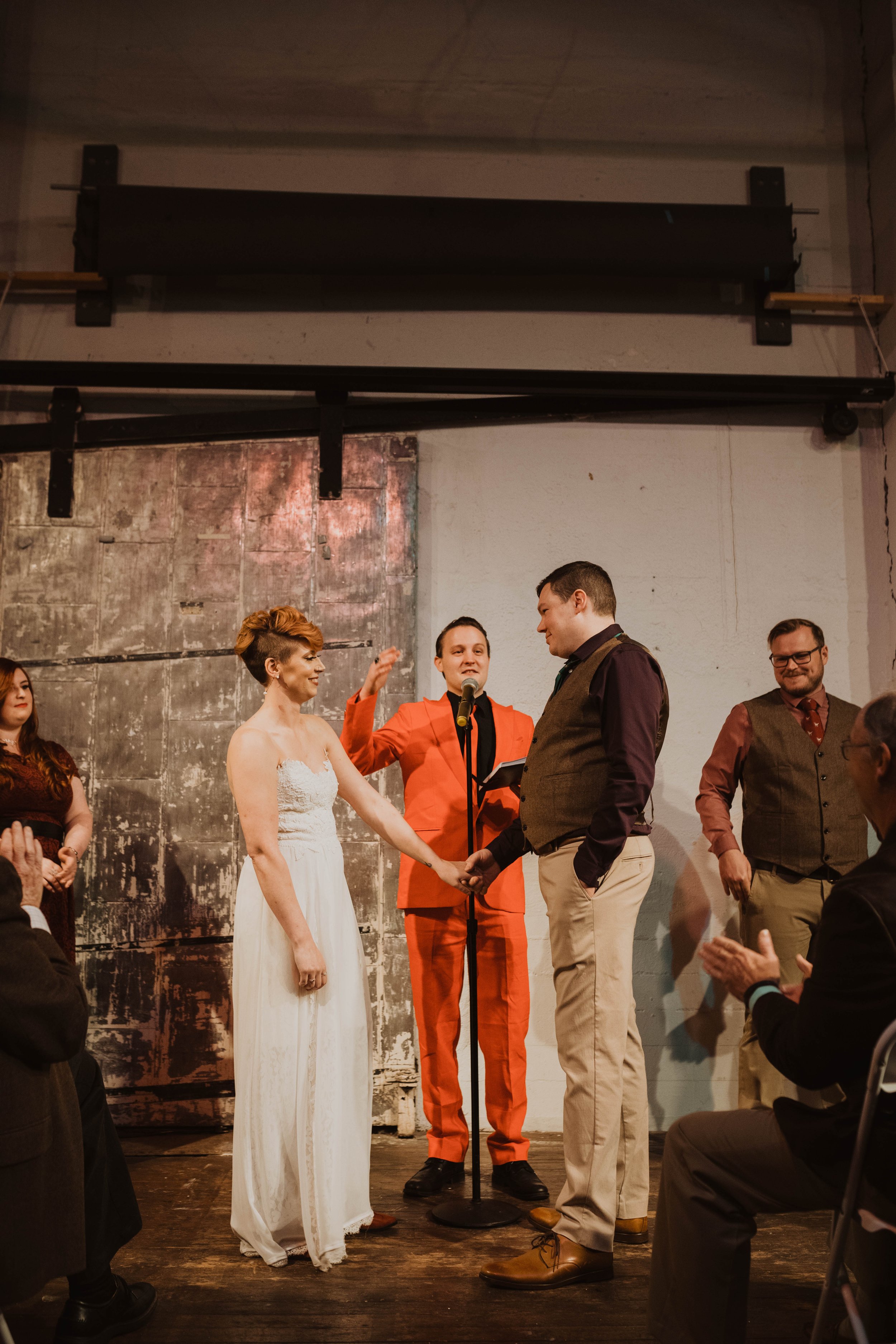 A wedding ceremony with a bride and groom exchanging vows on stage, surrounded by friends and officiant, with an audience clapping. Pioneer Square, Seattle, WA wedding photography.