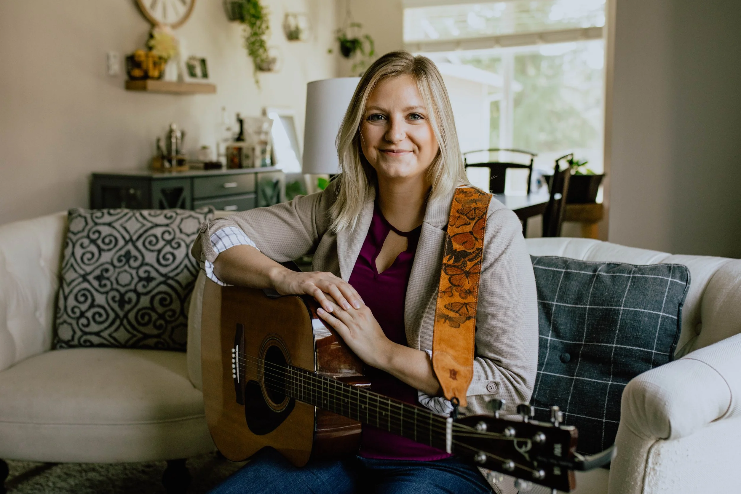 A woman with blonde hair smiling and sitting on a cream-colored sofa, holding an acoustic guitar with a butterfly-patterned strap. The room has a cozy, decorated living space with plants, photos, and a large window in the background.