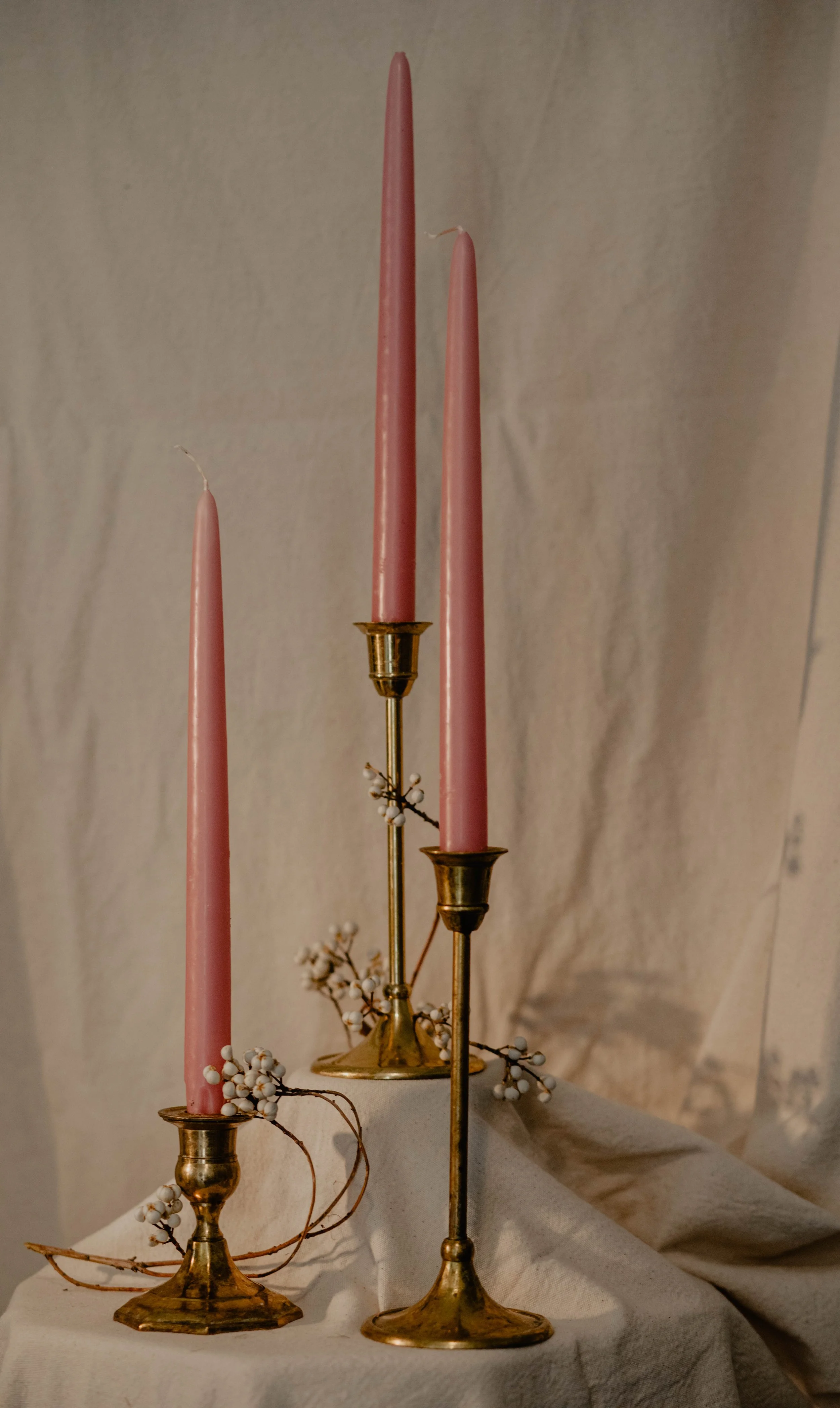 Three tall pink taper candles in vintage brass candlesticks, with small white berry branches halved on a white cloth, against a soft beige background.  Seattle product photography