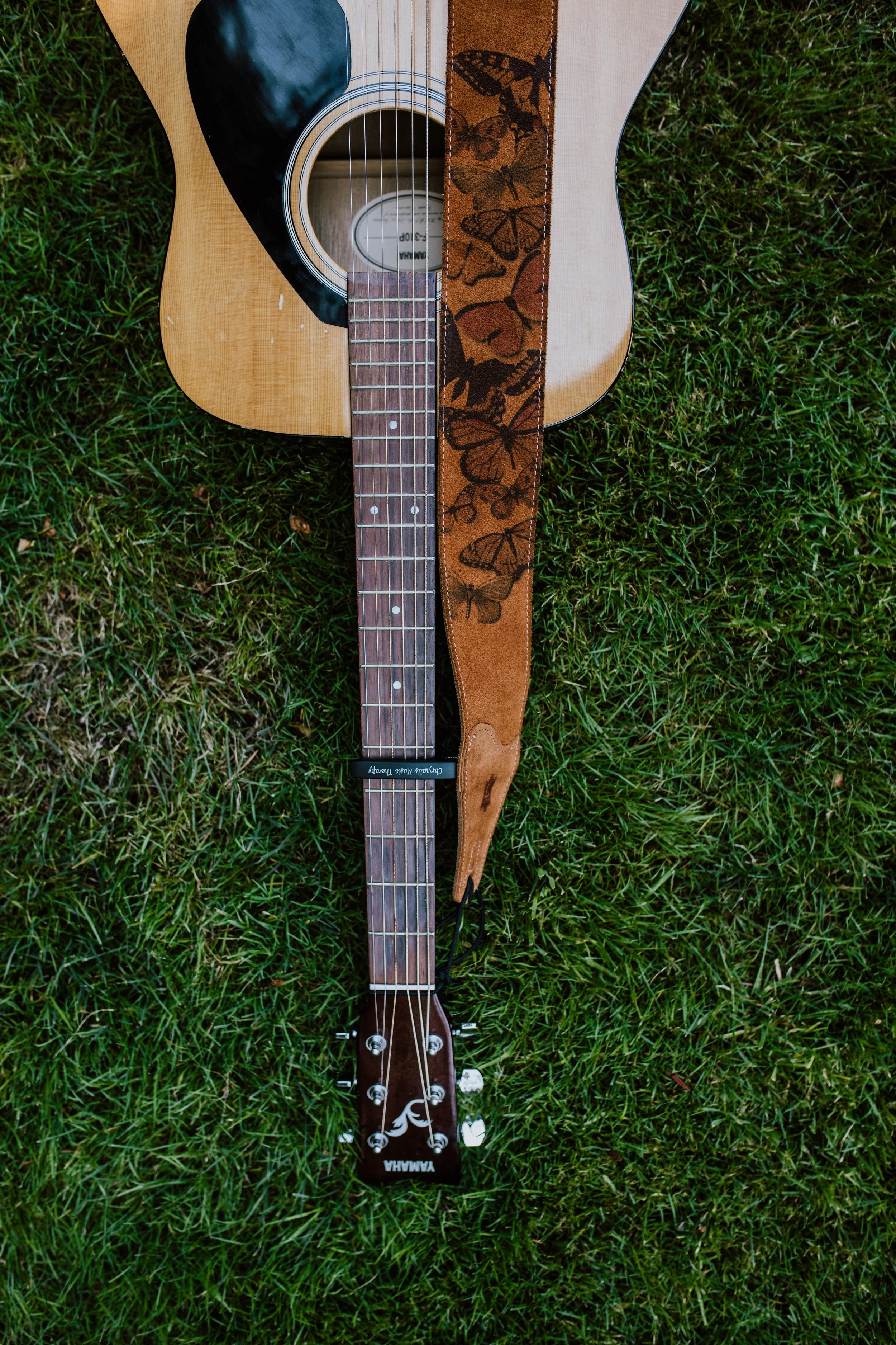 An acoustic Yamaha guitar with a brown strap adorned with butterfly designs lying on green grass. Seattle professional head shot photography