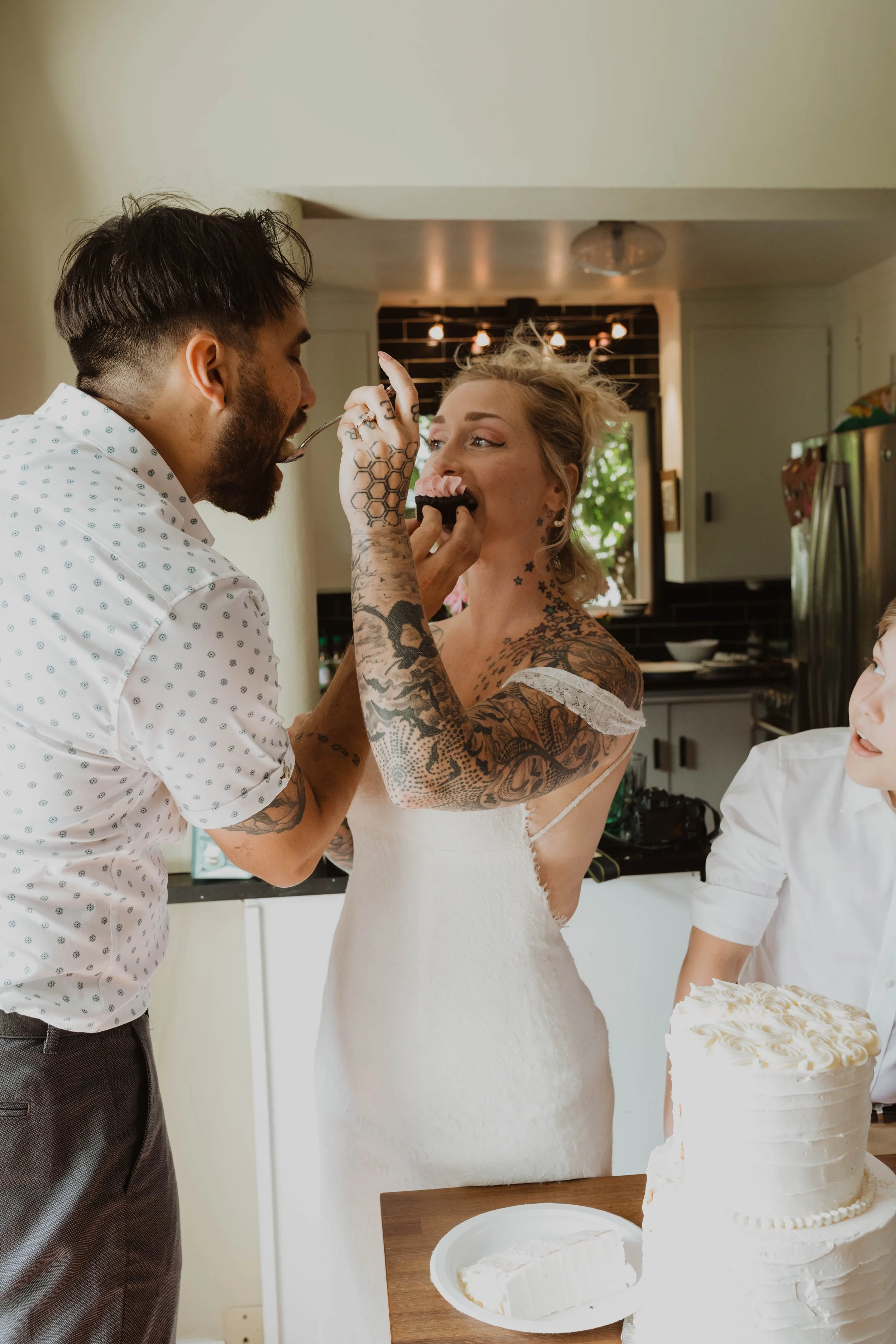 A woman in a wedding dress is being fed cake by a man, with a child looking on, in a kitchen setting during a celebration. Seattle, WA wedding photography.