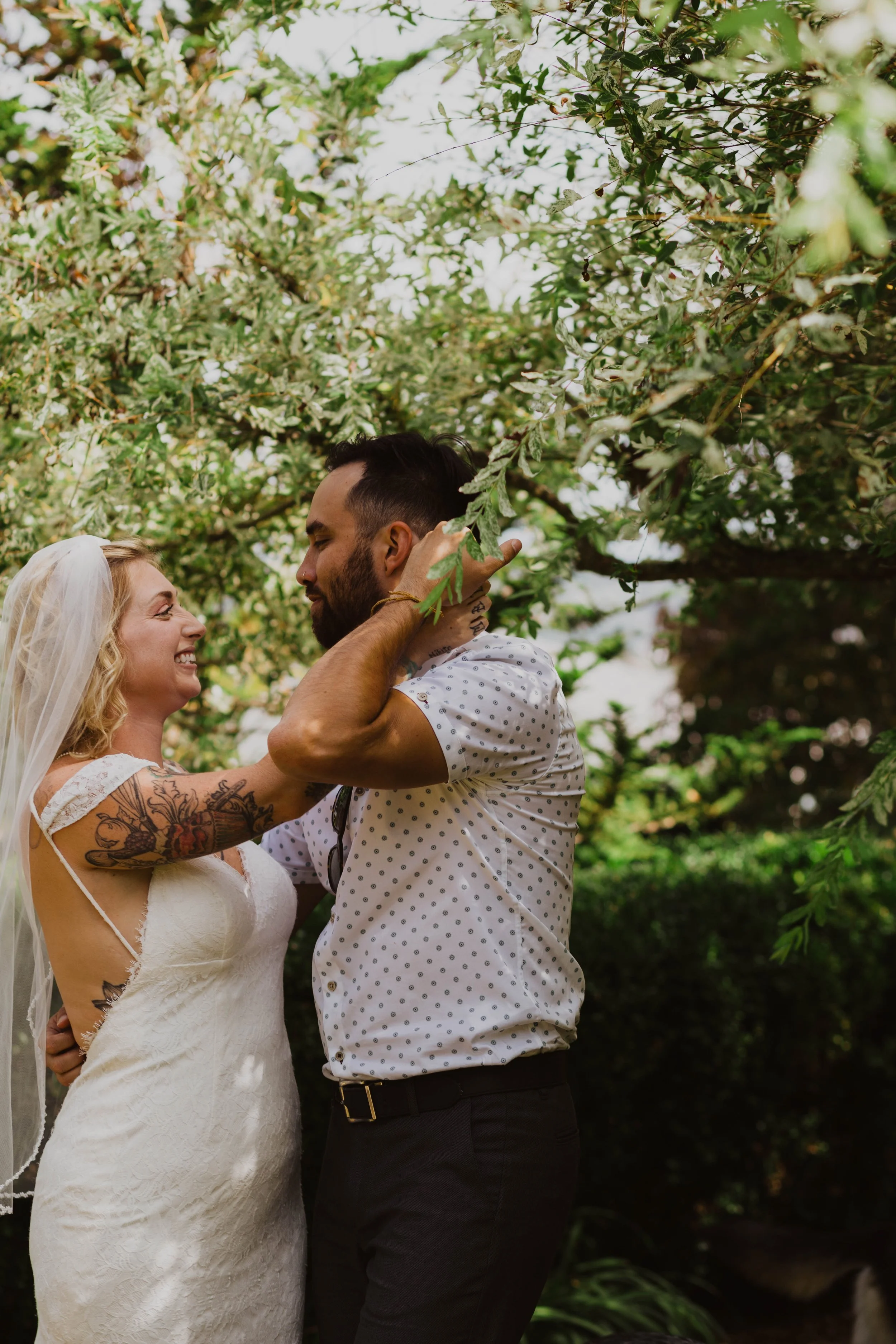 A happy couple with tattoos embrace in an outdoor setting surrounded by green leaves and trees, likely during their wedding or romantic moment. Seattle, WA wedding photography.