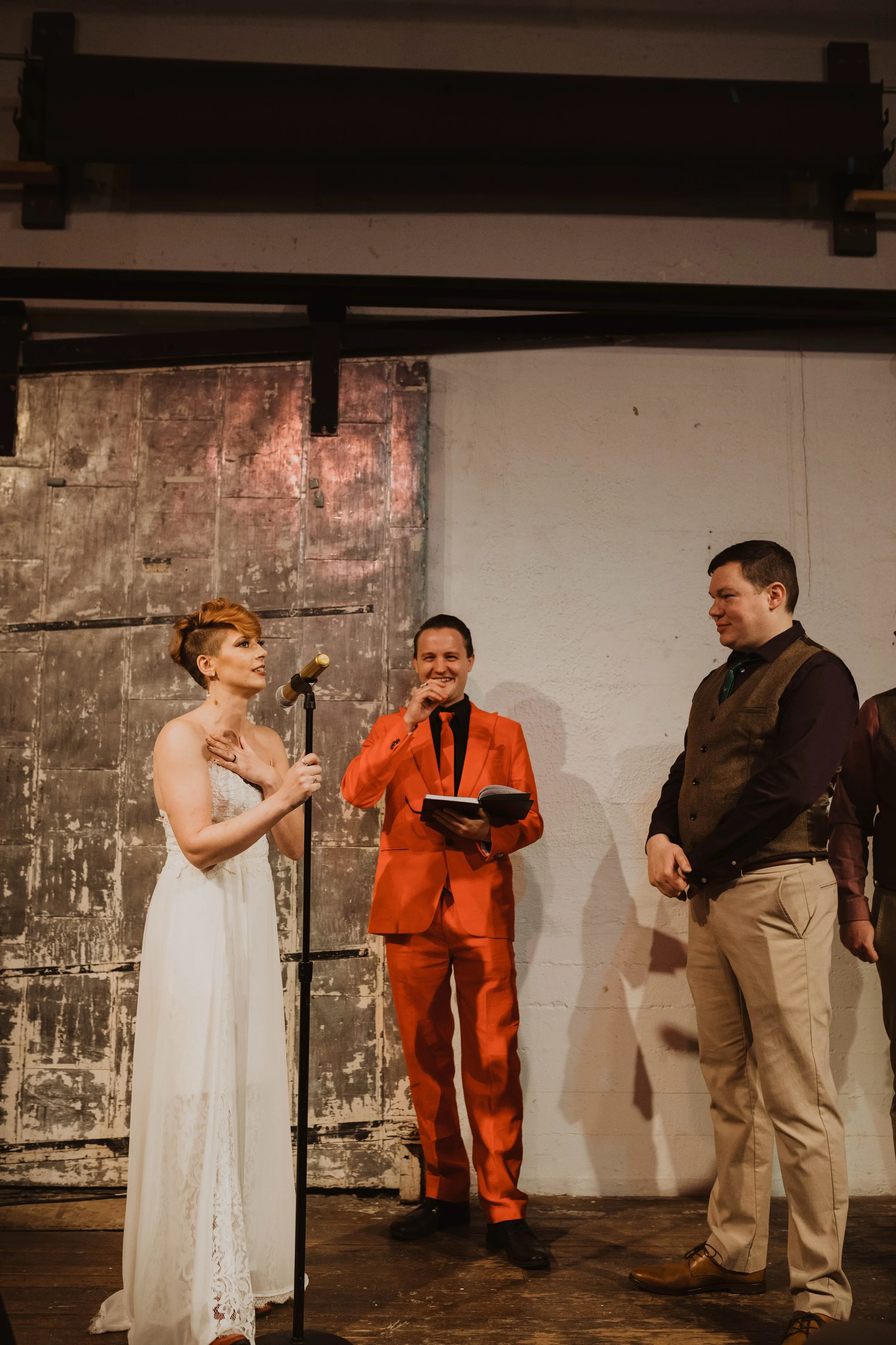 A woman in a white dress with short red hair reads from a microphone, two men listen, one in a bright orange suit with dark hair and another in a brown vest and beige pants, in an indoor setting with a rustic background.