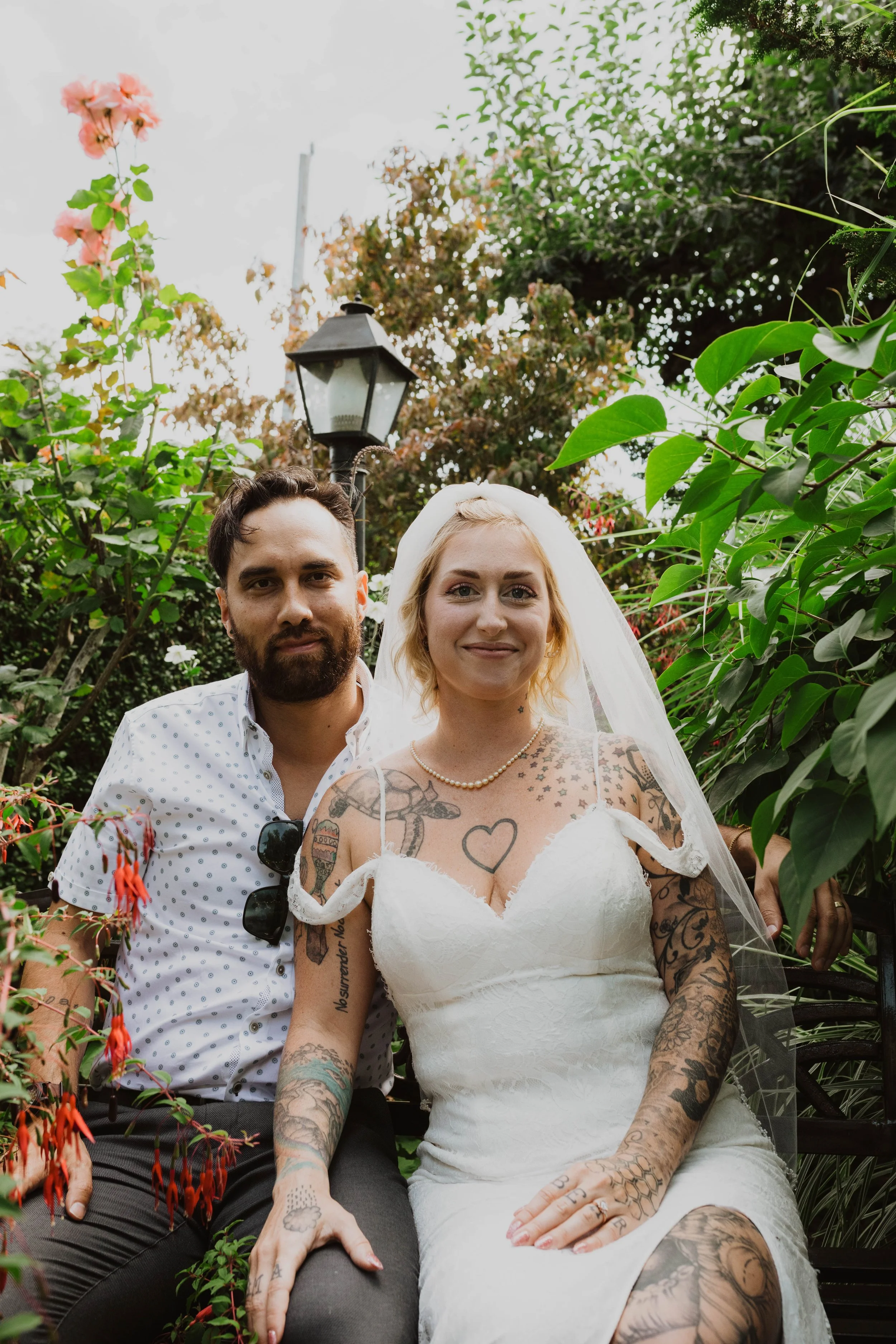 A newly married couple with tattoos, sitting outdoors on a garden bench, smiling at the camera, surrounded by green plants and flowering bushes, with a vintage street lamp in the background. Seattle, WA wedding photography.