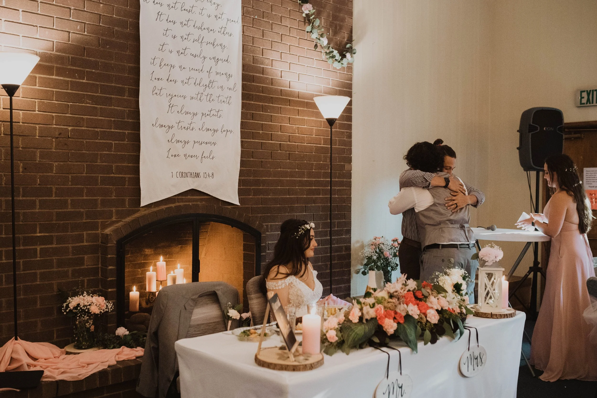 A wedding reception scene with a bride sitting at a decorated table with flowers and candles, a couple hugging near a microphone, and a woman reading or speaking at a table. There is a brick fireplace with candles and floral arrangements, and a large