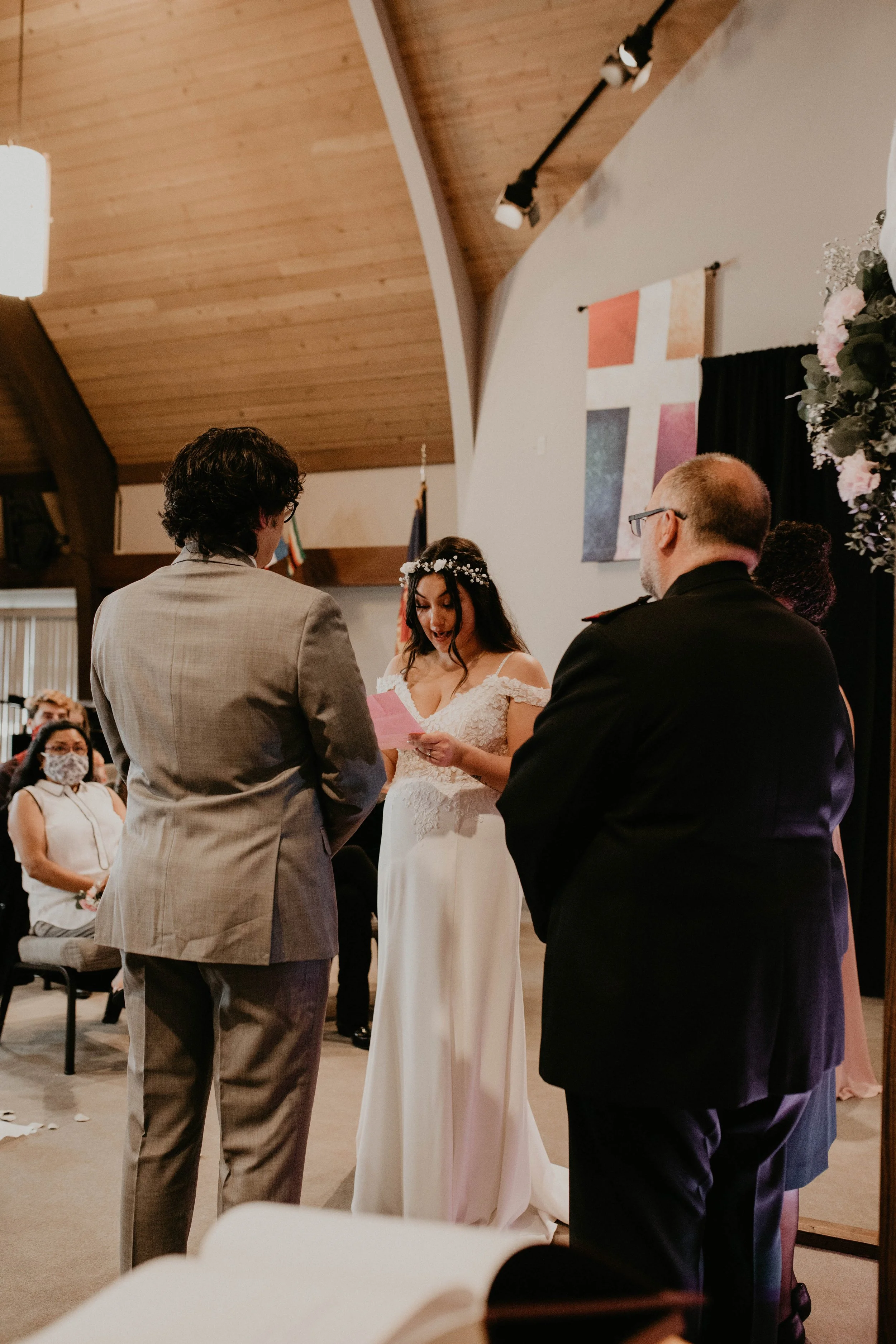 A bride with a floral headpiece and white wedding dress reads vows during a wedding ceremony, with the groom and officiant in front of her in a church or chapel setting. Seattle, WA wedding photography.