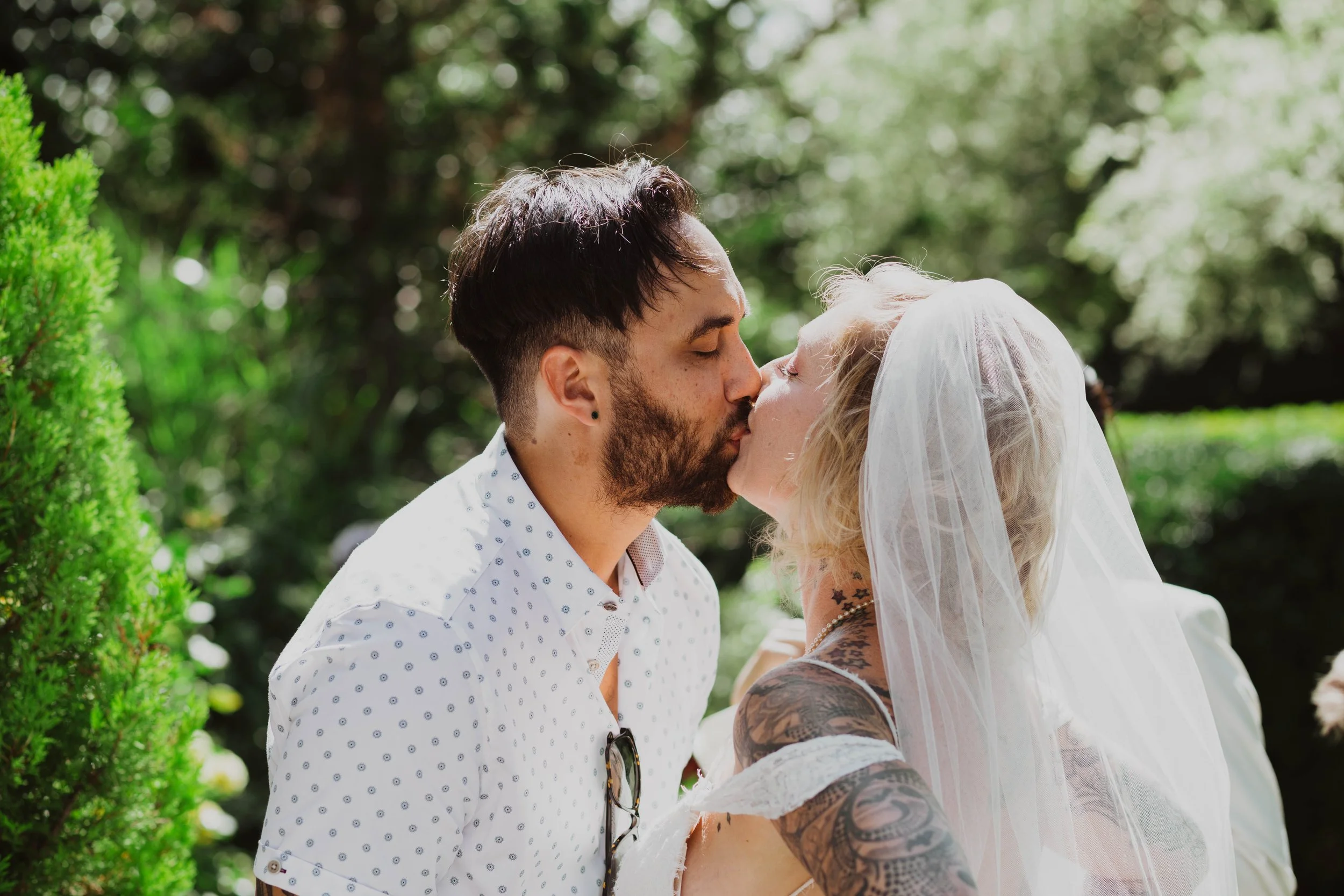 A man and woman kiss outdoors, with the woman wearing a white veil and having tattoos on her arms, and the man wearing a white shirt with small blue patterns. Seattle, WA wedding photography.