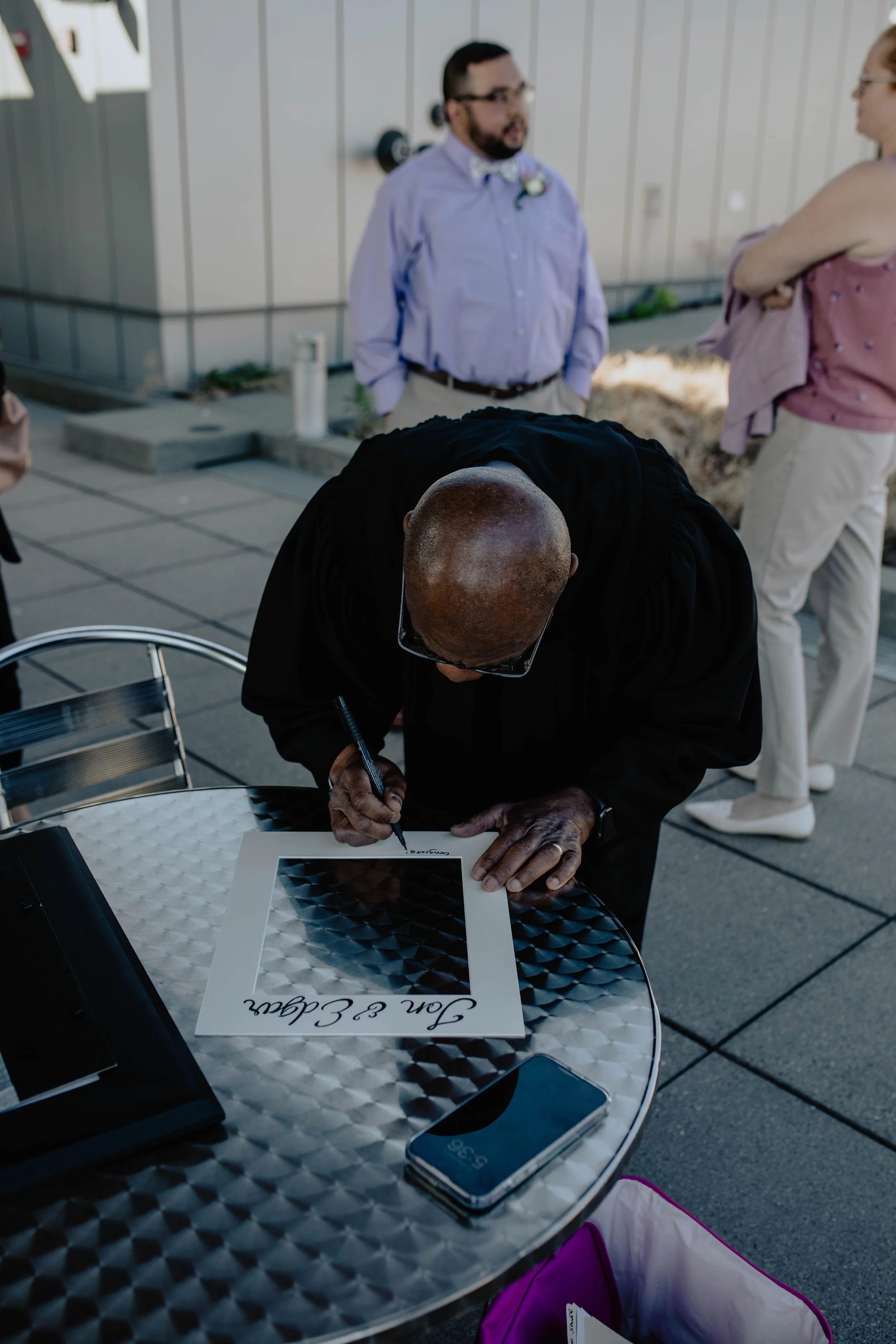Man signing a poster with a geometric black and gray pattern and the words 'Can & Caan' at the bottom, at an outdoor event with other people in the background. Seattle Municipal Courthouse wedding photography.