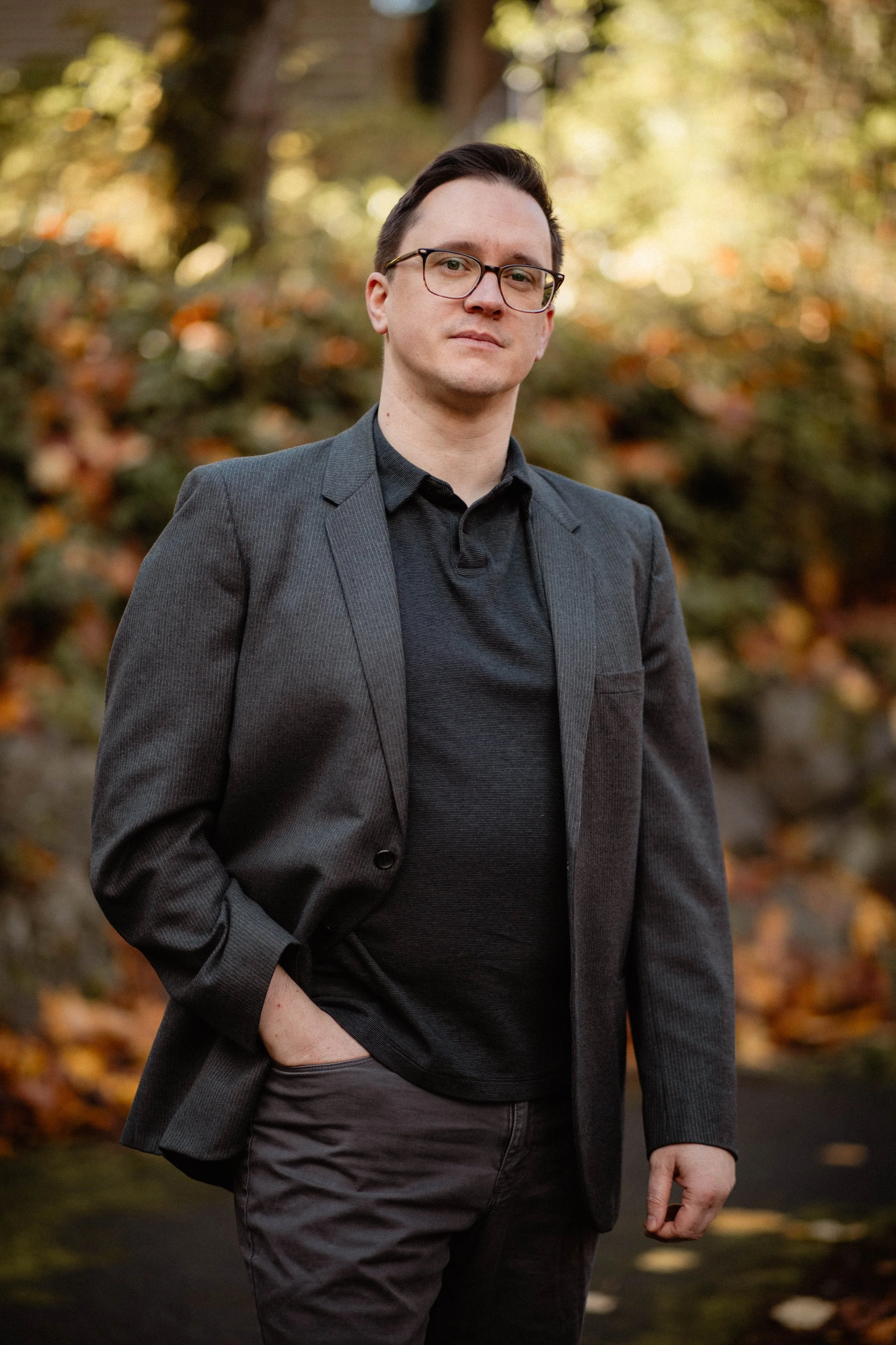 A man in a gray blazer and black shirt standing outdoors with autumn foliage in the background. Seattle professional head shot photography