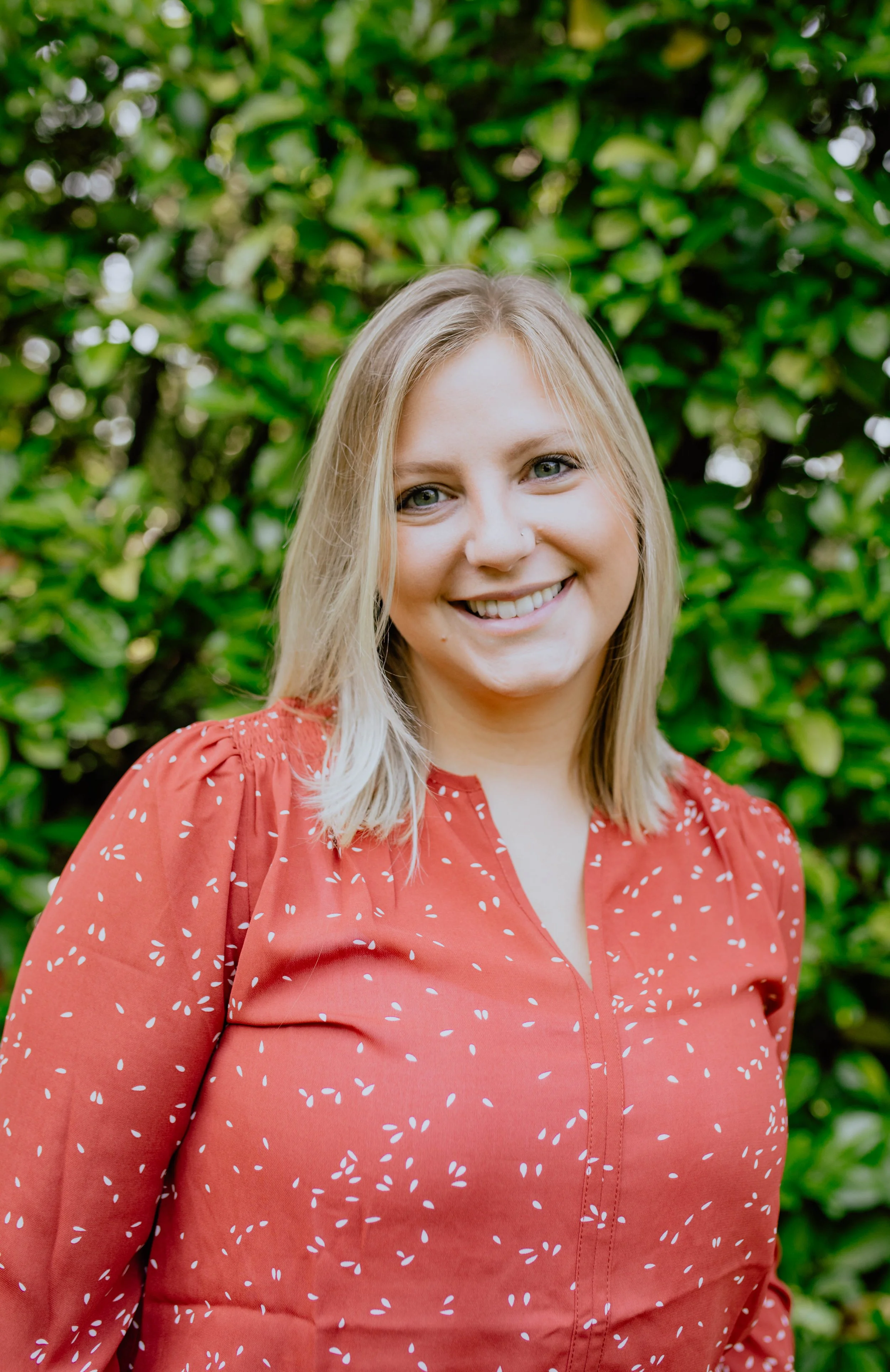A young woman with blonde hair and a nose piercing, smiling, wearing a red blouse with white patterns, standing outdoors in front of green leafy bushes. Seattle professional head shot photography