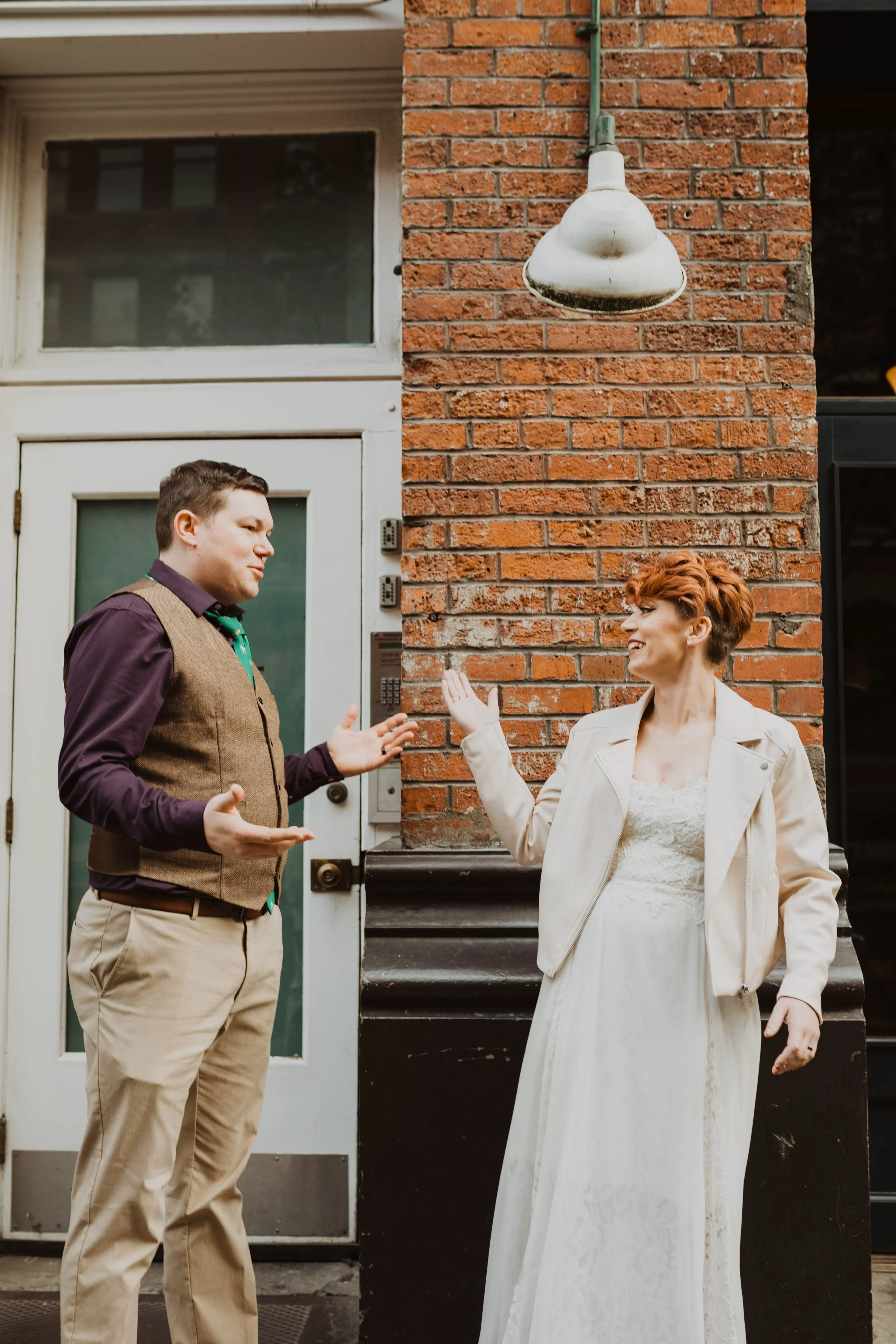 A man and a woman are standing outside a building, engaging in a conversation. The man, wearing a brown vest over a purple shirt and beige pants, is gesturing with his hands. The woman, with short red hair, wearing a white dress and a light-colored j