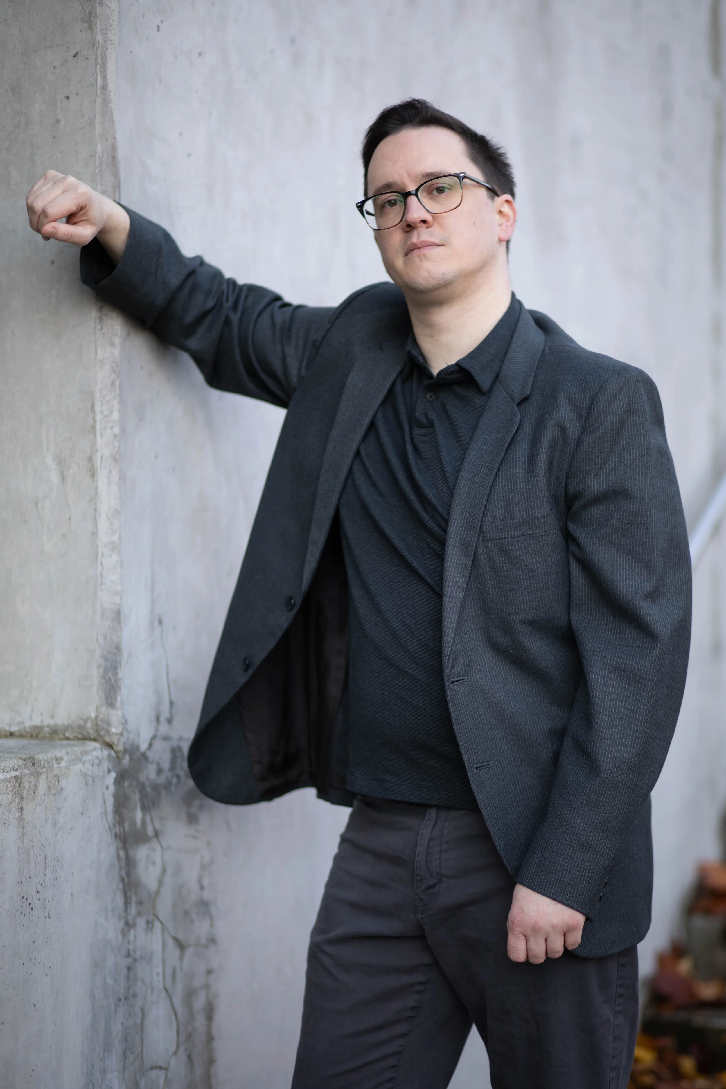 Man in black shirt and gray blazer leaning against a concrete wall with a serious expression. Seattle professional head shot photography
