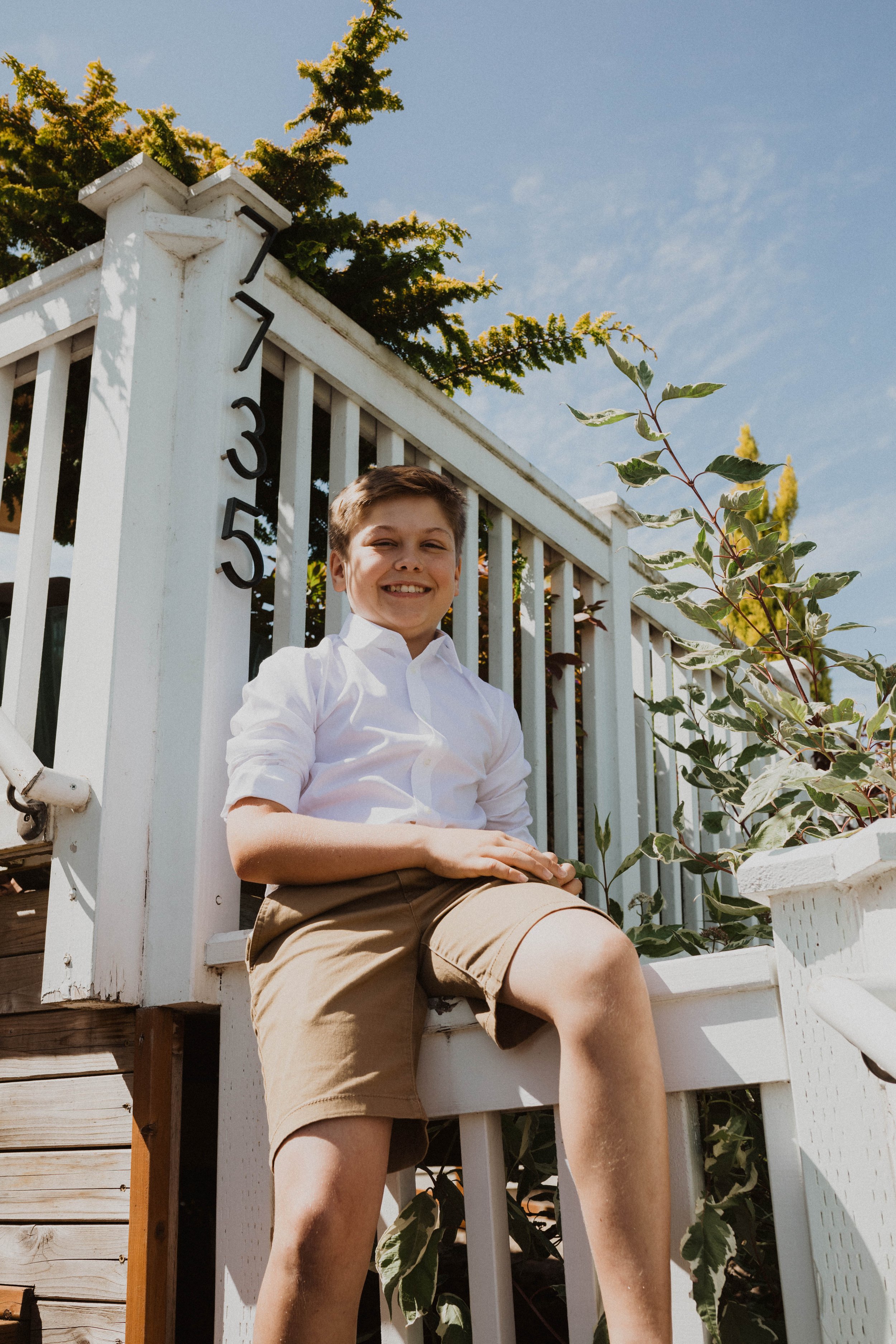A smiling boy in a white shirt and khaki shorts sitting on the white porch railing of a house. The house has the number 735 on the post, with blue sky and trees in the background. Seattle, WA wedding photography.