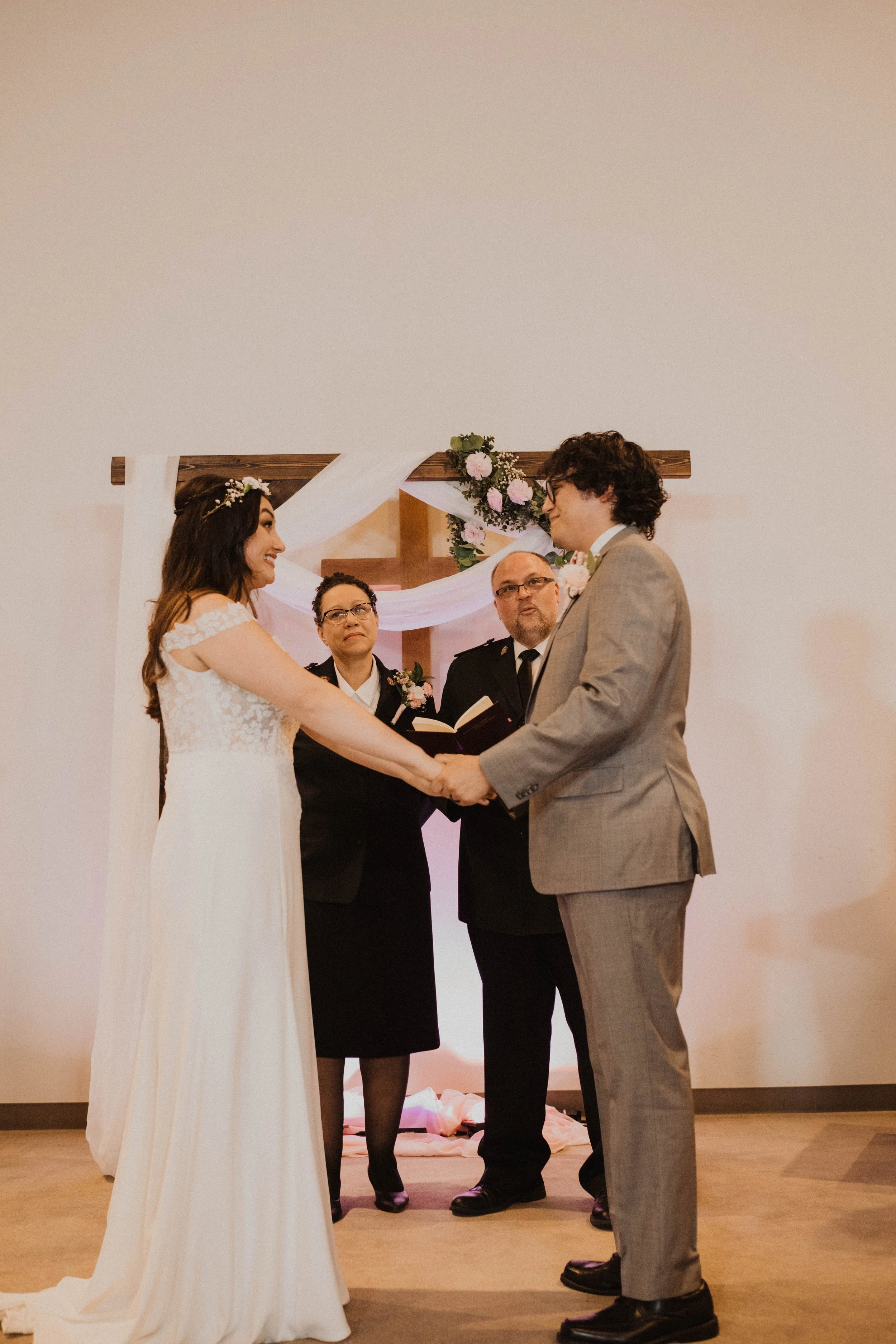 A couple getting married, holding hands and facing each other, in a wedding ceremony. The bride is wearing a white wedding dress and the groom is in a gray suit. Two officiants, one women and one man, stand behind them, officiating the ceremony under