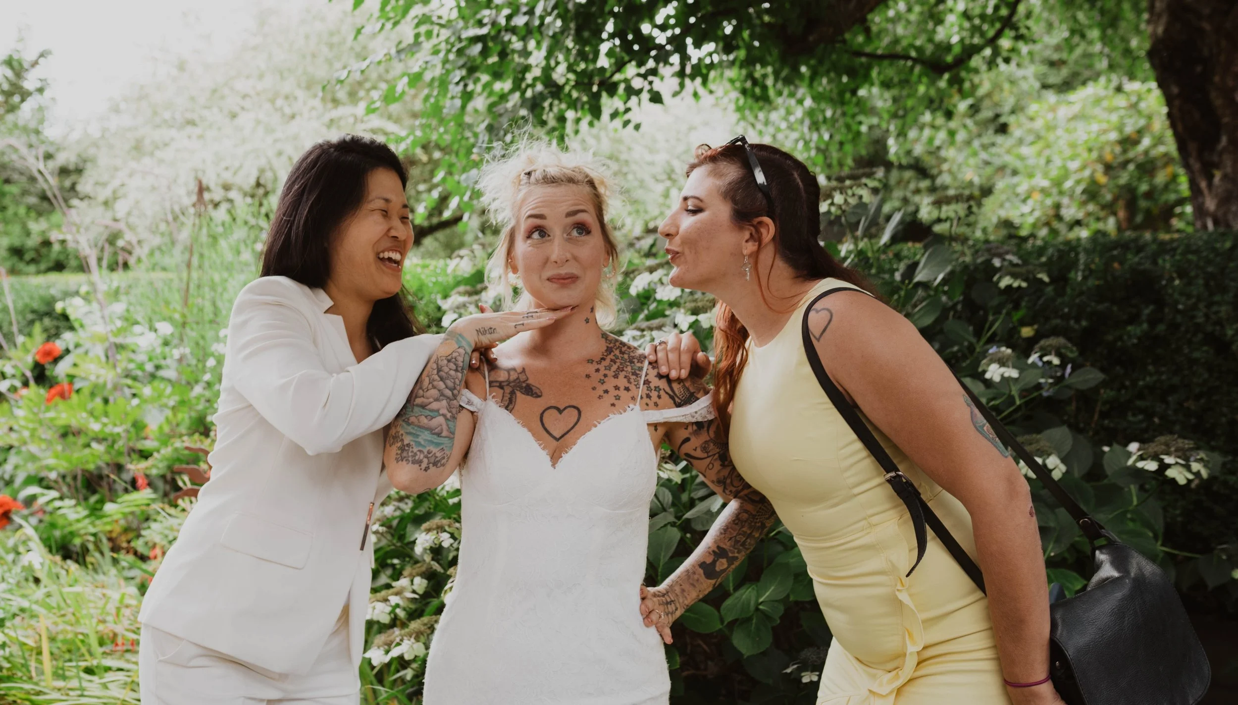 Three women, one in a wedding dress, posing playfully with expressions, outdoors in a garden with green foliage and flowers. Seattle, WA wedding photography.