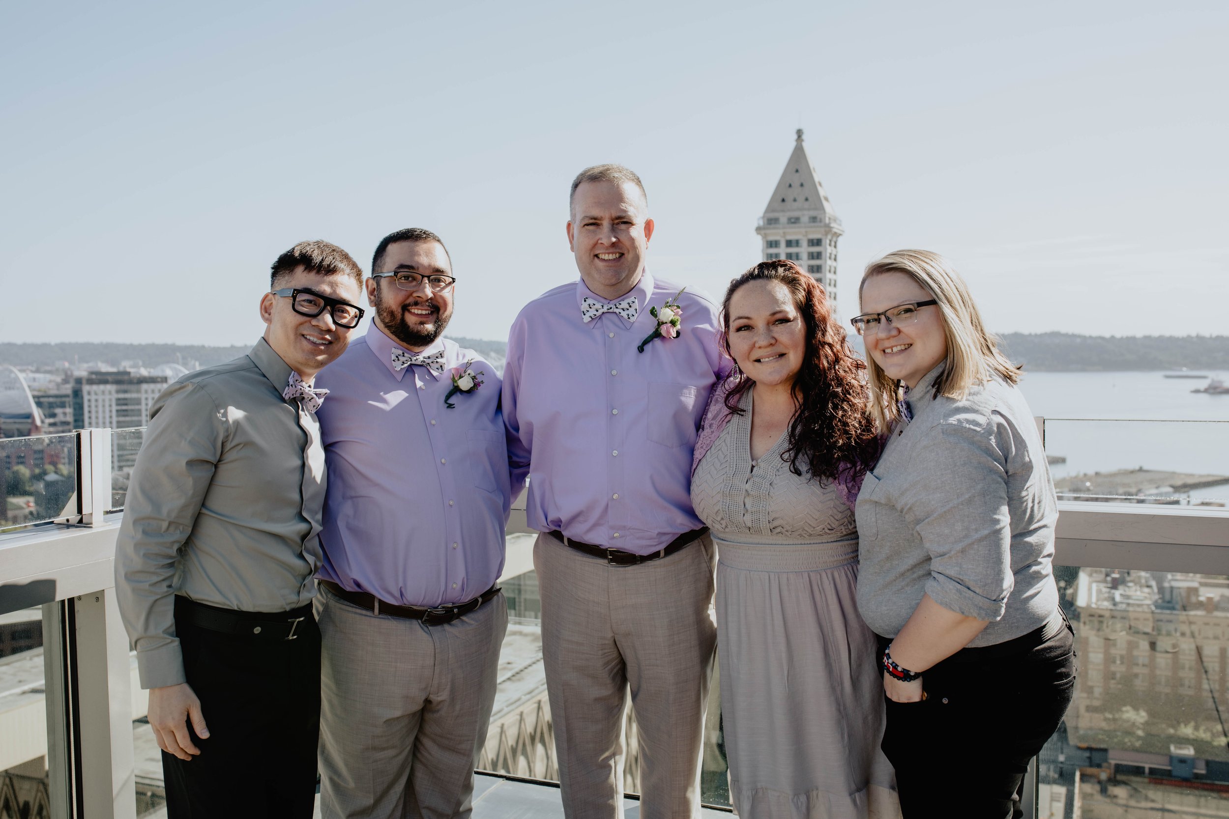 Group of six people standing on a balcony outdoors, with a cityscape and water in the background, smiling at the camera, dressed in semi-formal attire with some wearing bow ties. Seattle Municipal Courthouse wedding photography.