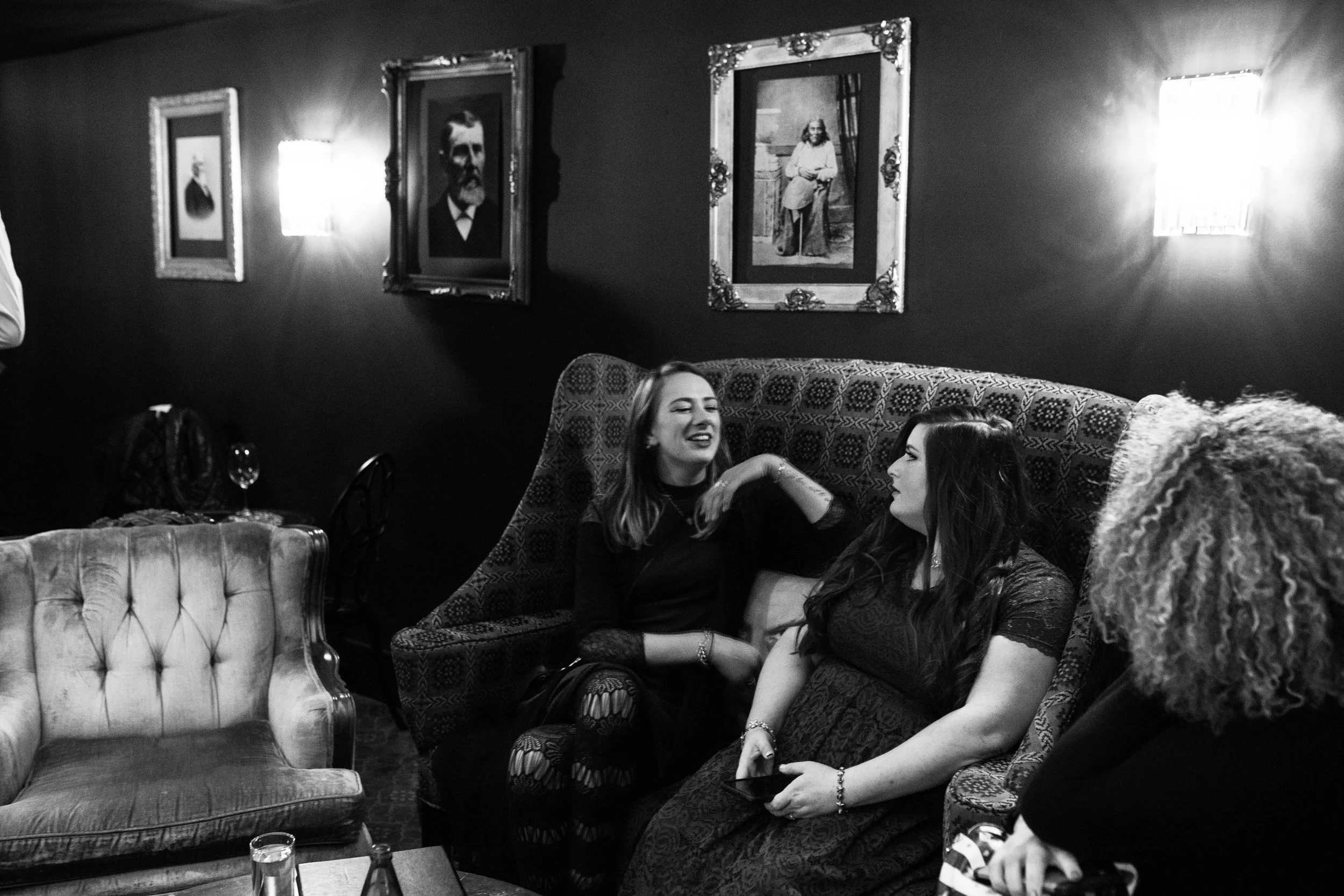 Group of women sitting and chatting on a vintage sofa in a dimly lit room decorated with framed portraits, with a table in front holding glasses and bottles. Pioneer Square, Seattle, WA wedding photography.