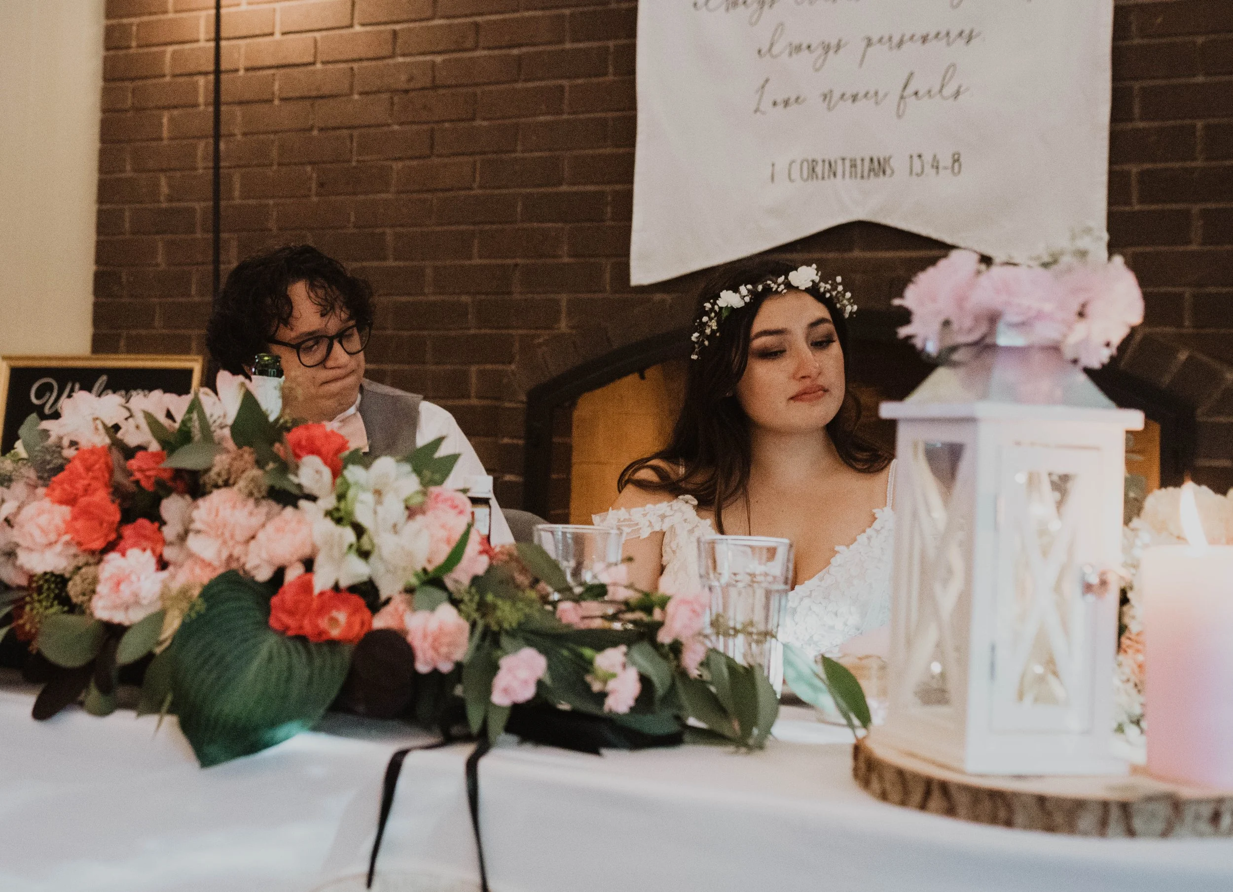 At a wedding reception, a woman in a white dress with a floral headband sits beside a man with glasses and curly hair. The table is decorated with pink, red, and white flowers, green leaves, a white lantern, and a pink candle. A large banner with rel